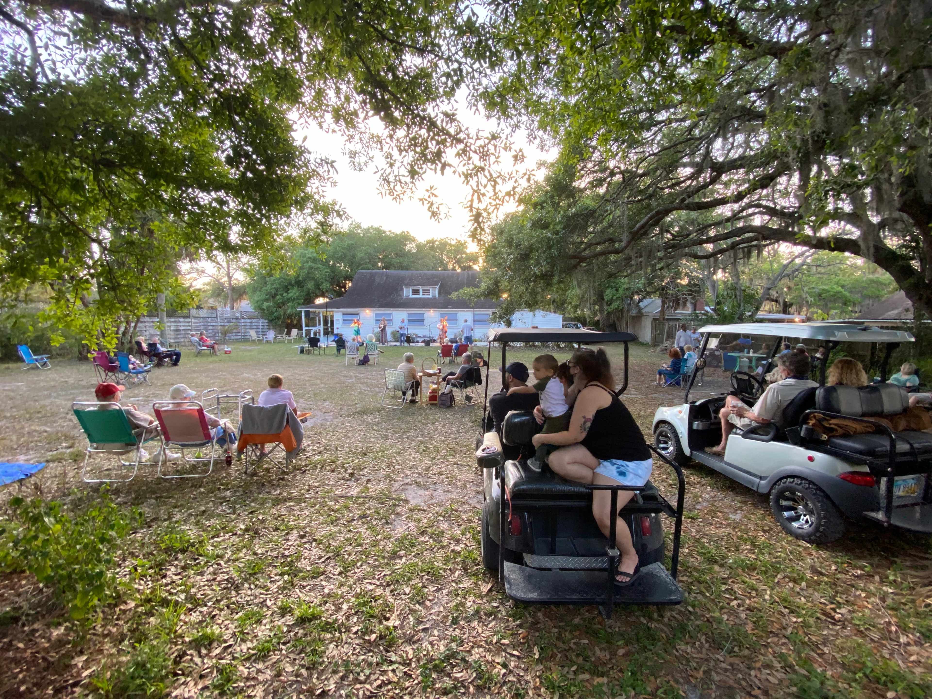 A group of people gather in a grassy area with golf carts parked nearby, while others sit in chairs facing a house in the background.