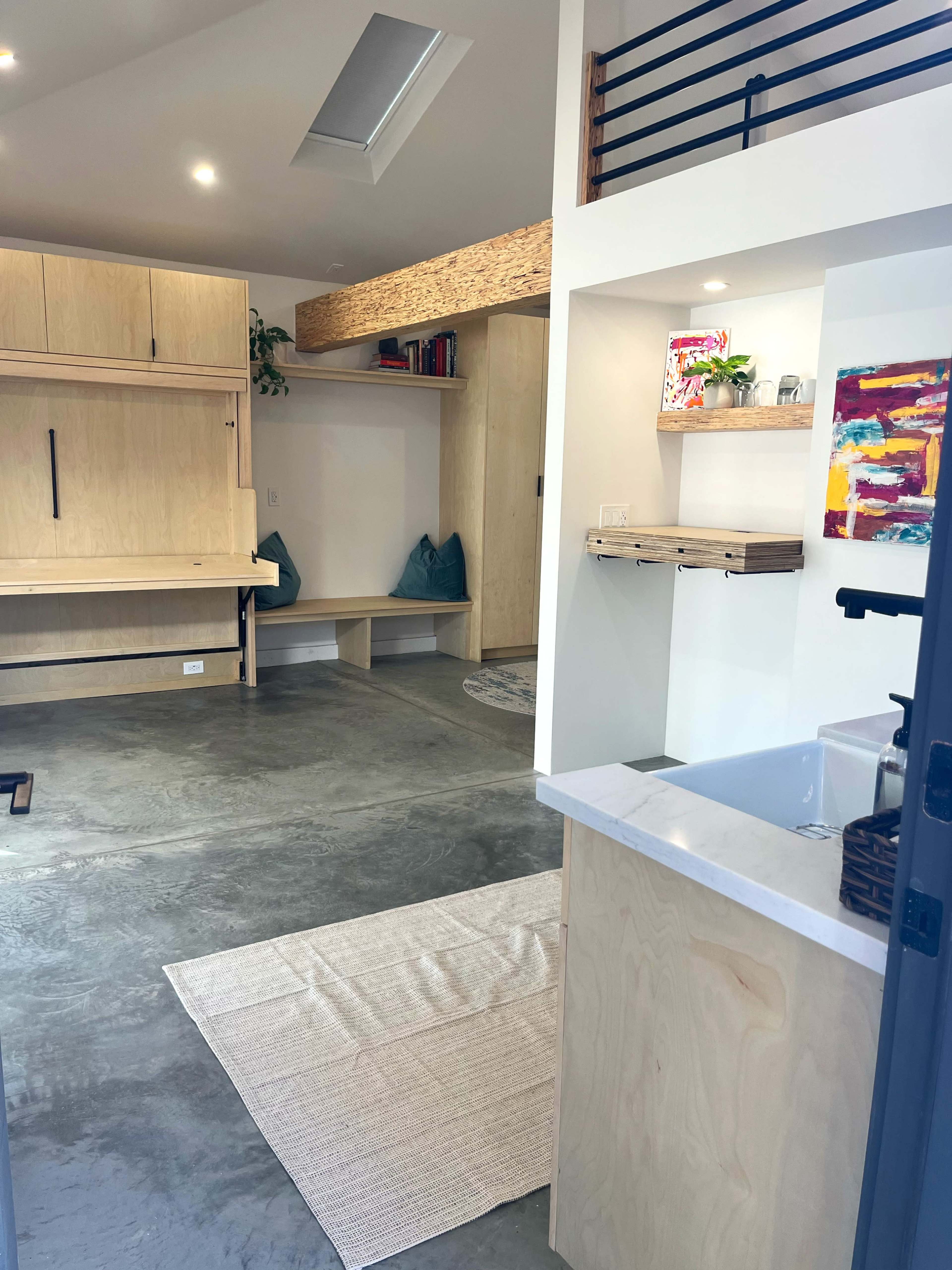 The image shows a modern interior space featuring a minimalist design with wooden storage units, a small kitchen area, concrete flooring, and a cozy seating nook.