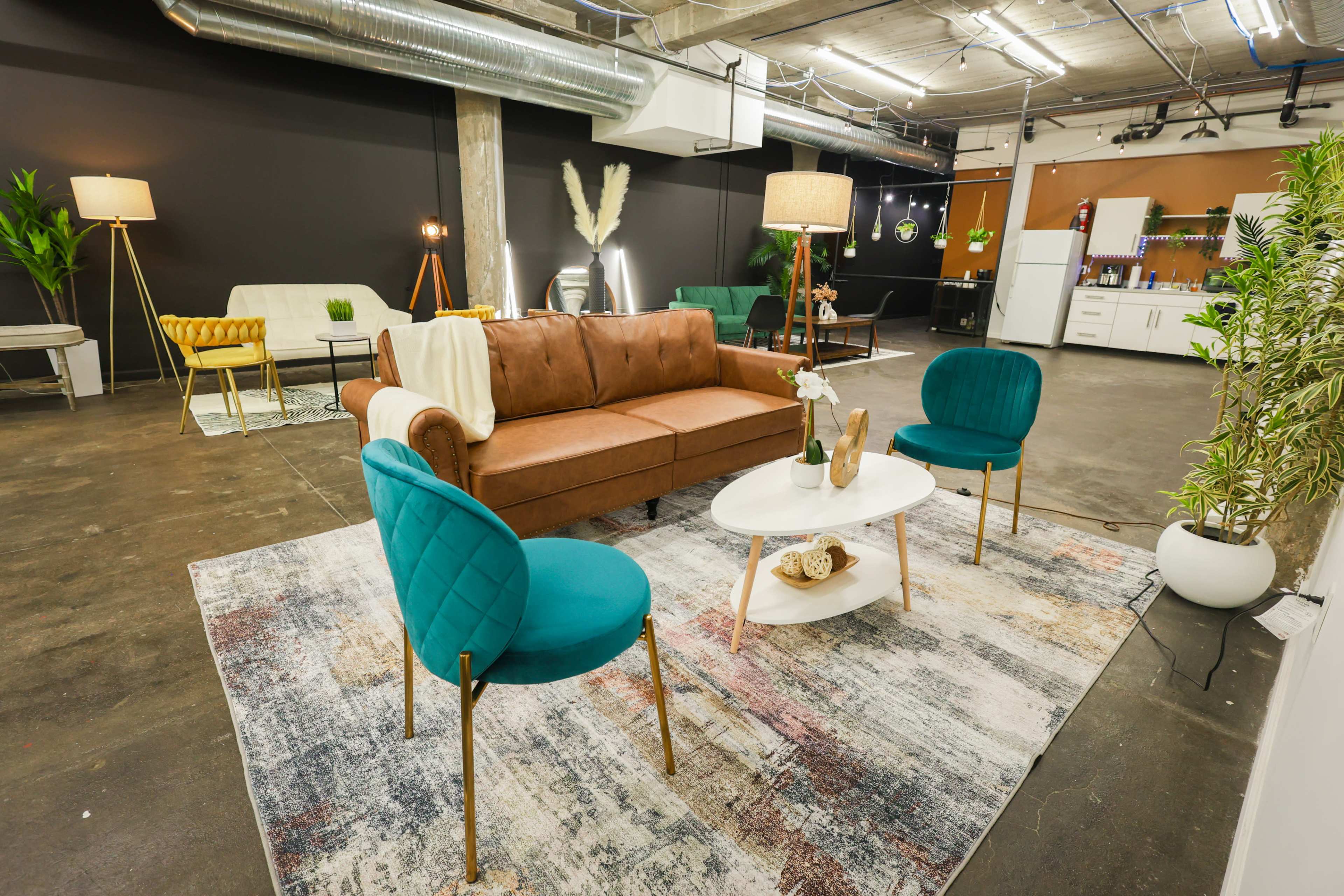 A modern interior space featuring a brown leather sofa, four teal chairs, a round white coffee table, and various plants, with an open kitchen area in the background.