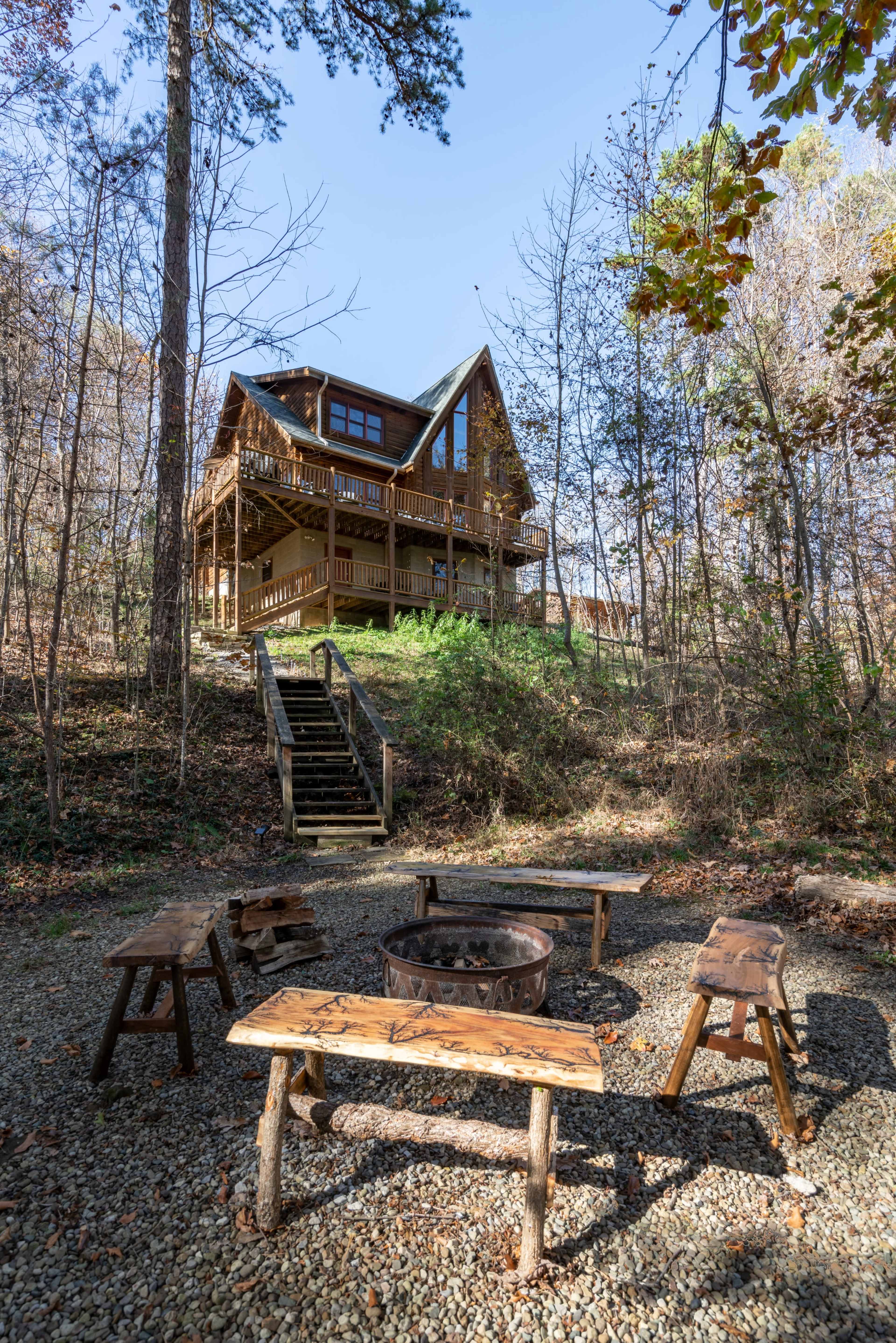 A wooden cabin with a steep roof stands on a hillside, surrounded by trees, with a fire pit and benches arranged in front of it.