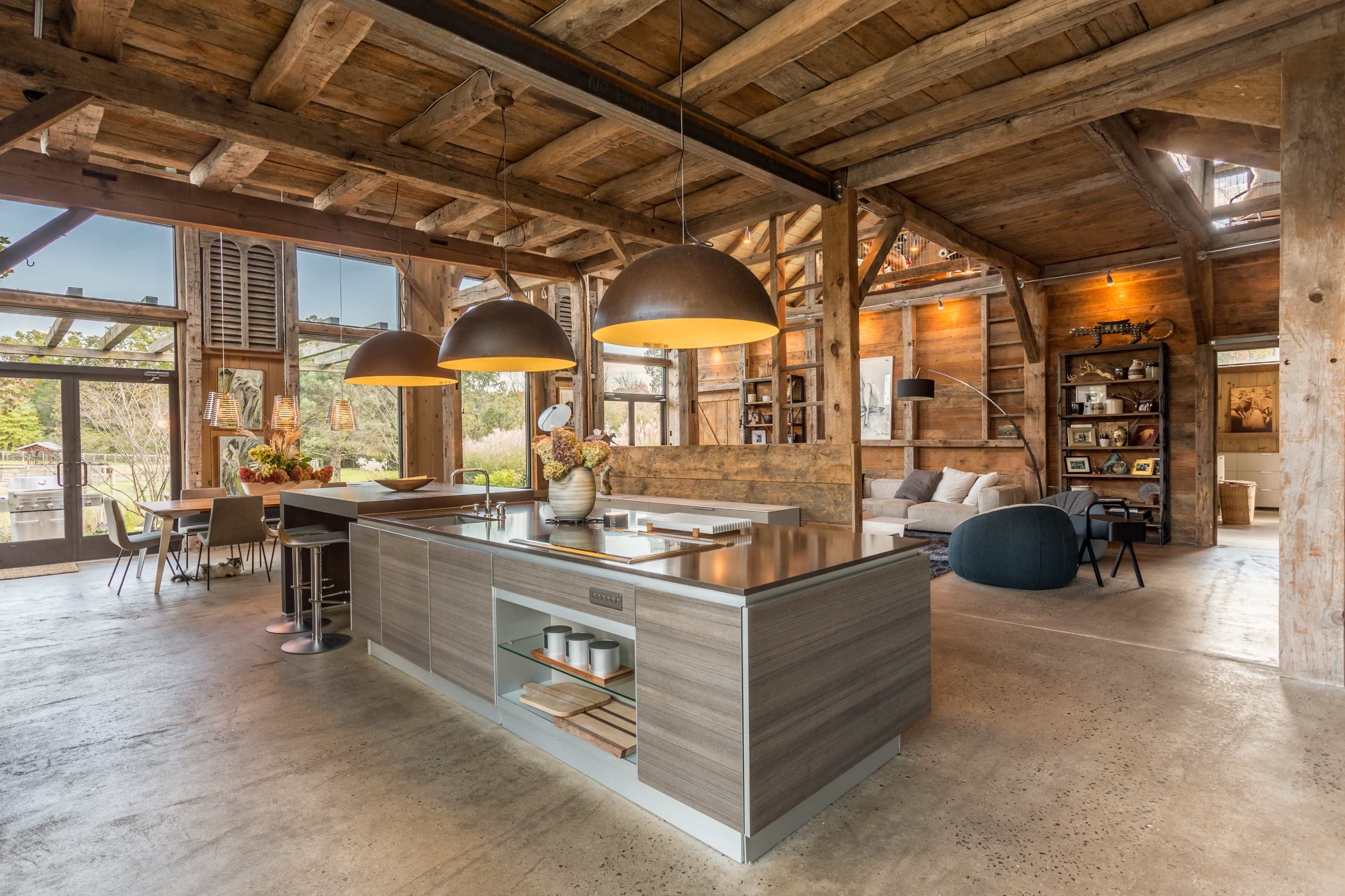 A modern kitchen with a central island, wooden beams, and large pendant lights, set in a rustic interior with large windows.
