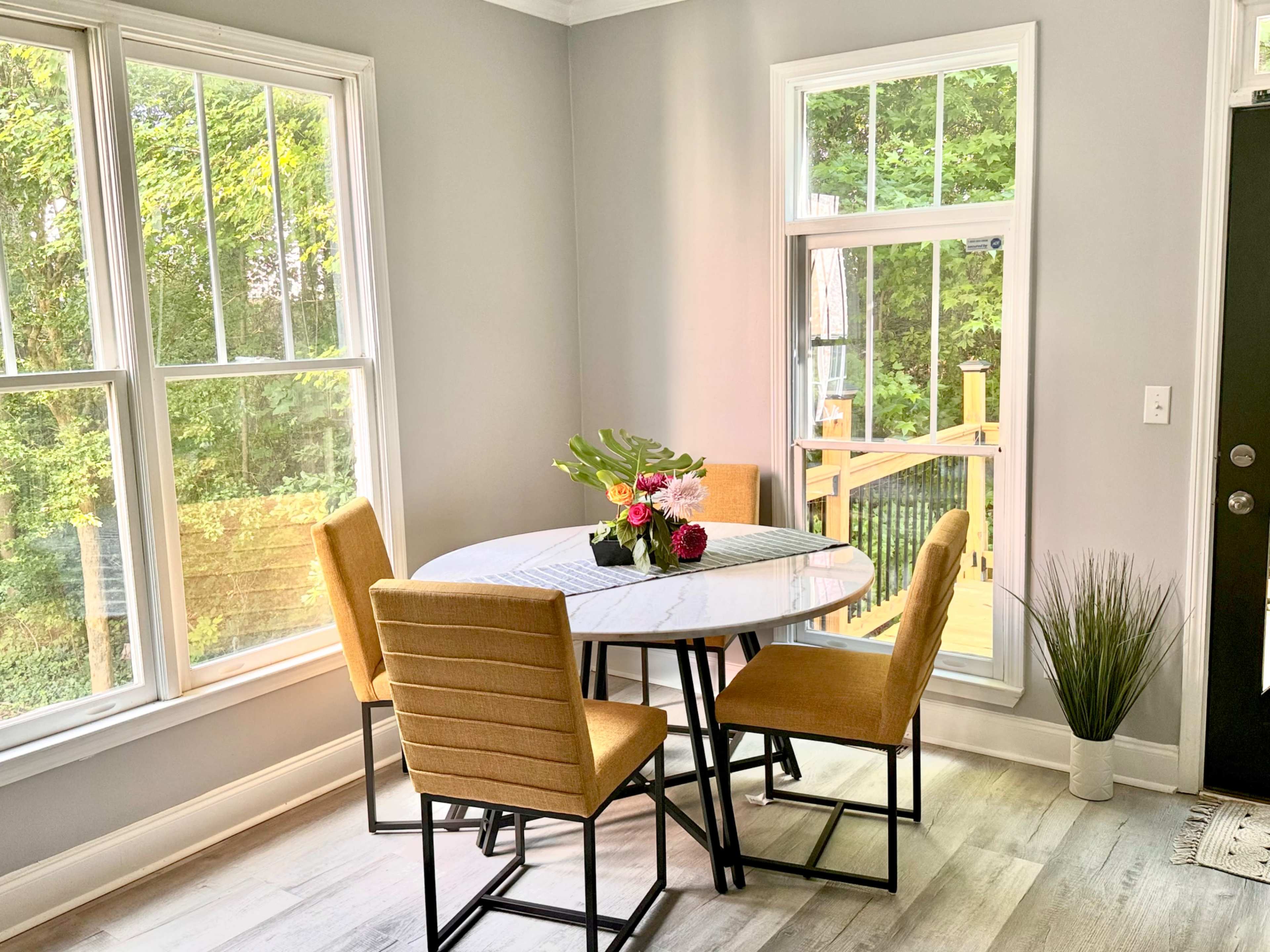 A round dining table surrounded by four mustard-colored chairs is set near large windows that offer a view of greenery outside.