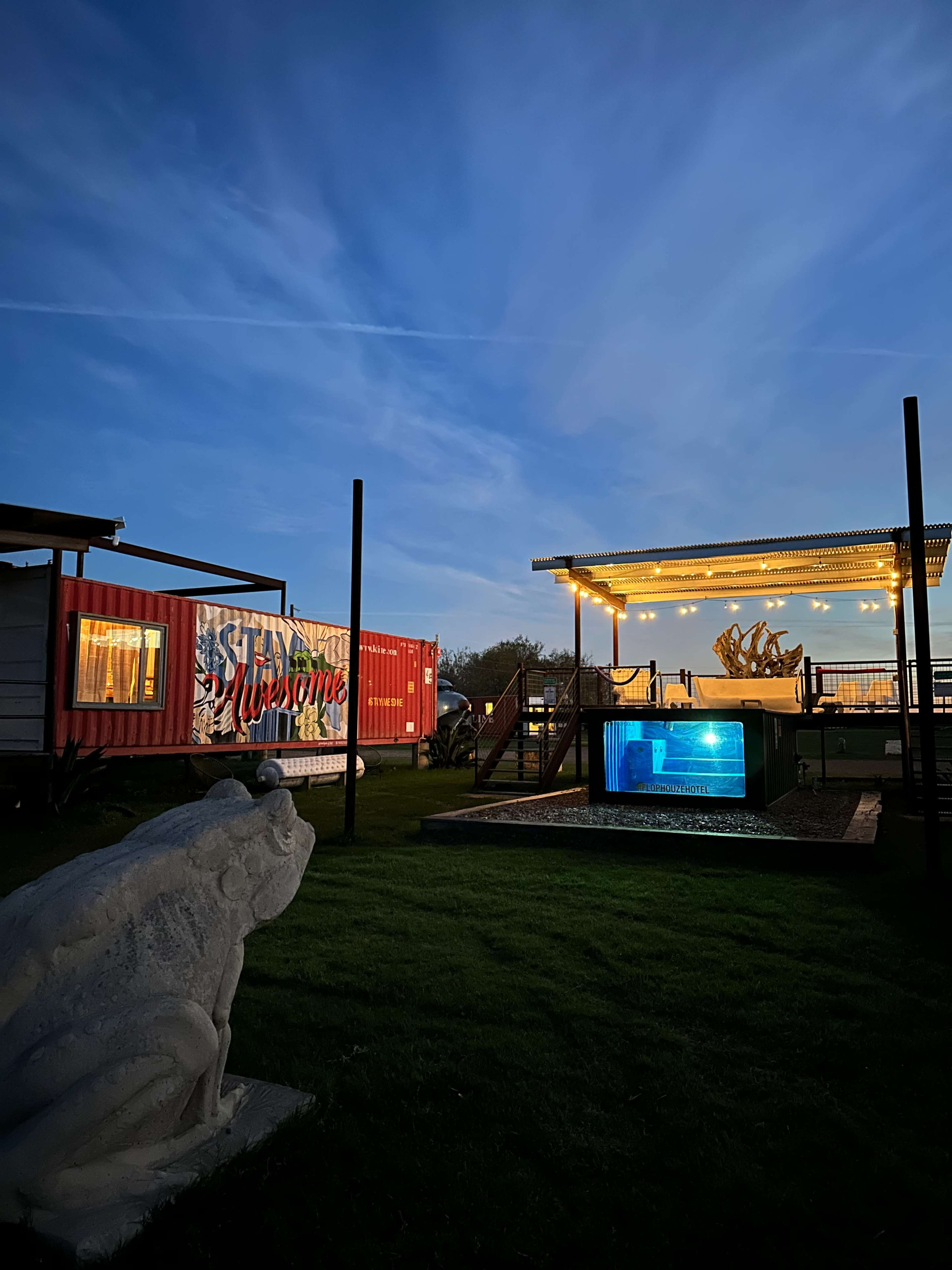 A colorful shipping container marked "Welcome," a well-lit patio with decorative lights, and a stone frog sculpture in a grassy area during twilight.