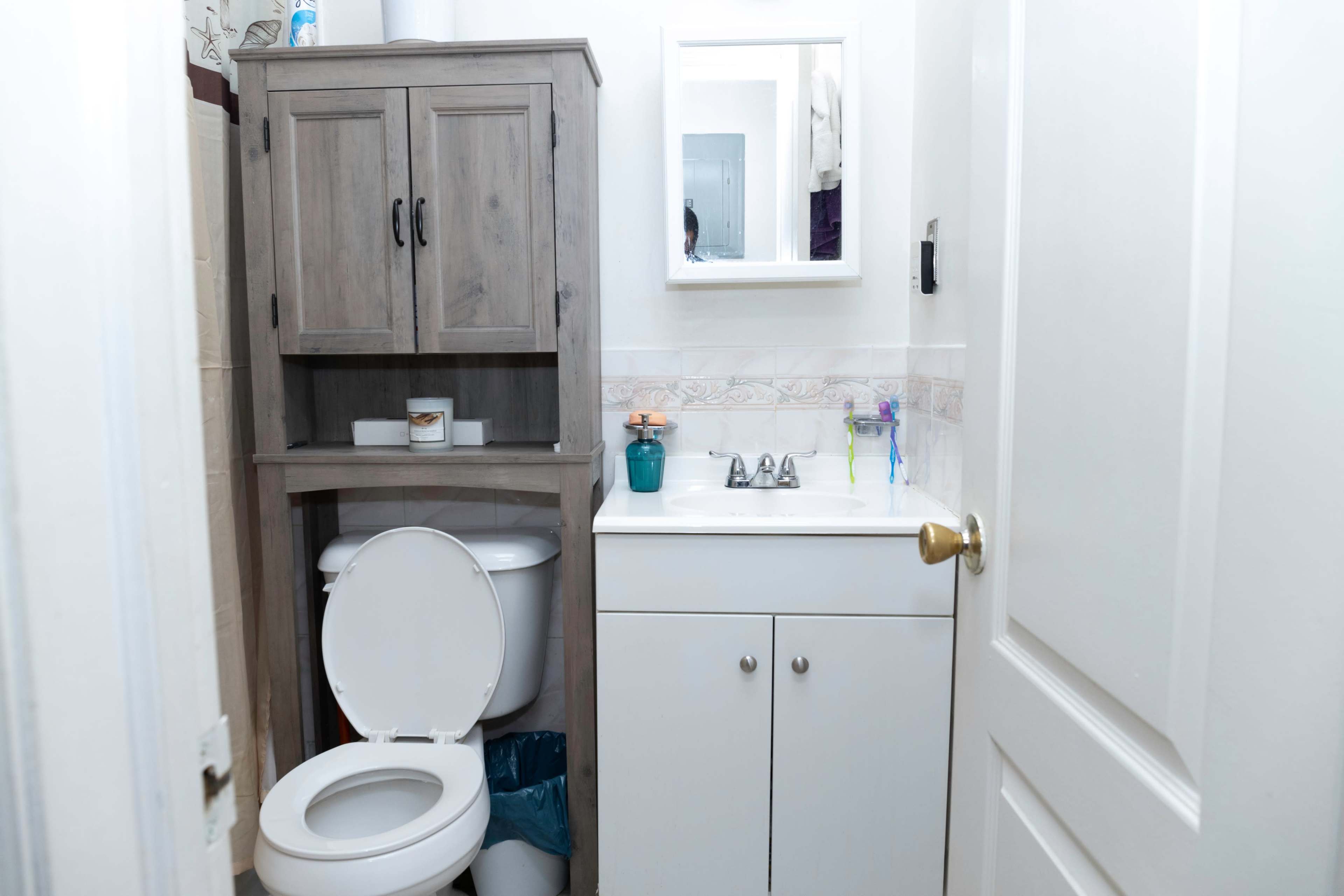 A small bathroom featuring a toilet, a sink with a cabinet underneath, and a mirror above the sink, with a storage unit beside the toilet.