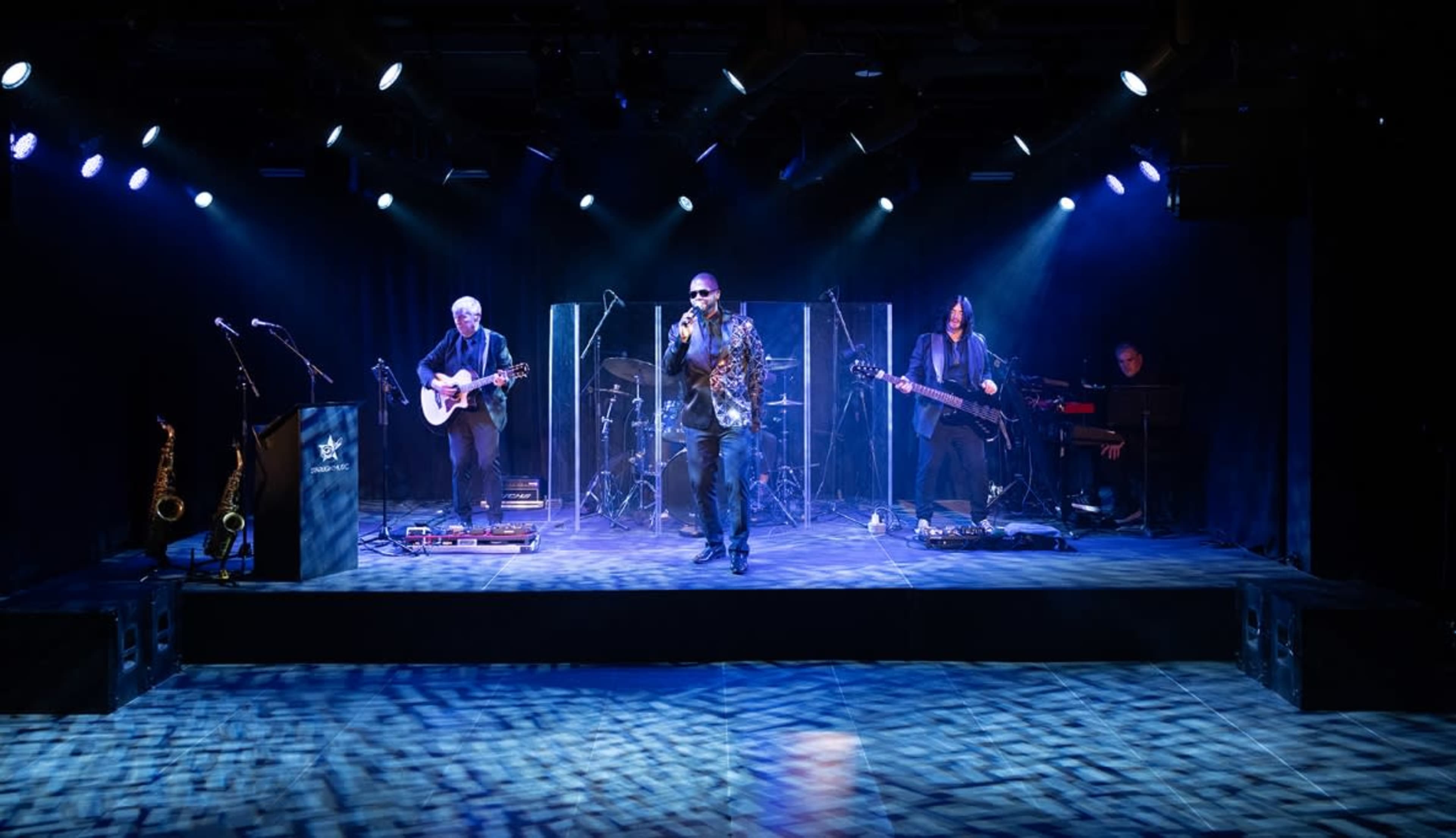 A live band performs on stage with illuminated instruments and a vocalist in front of a transparent screen.