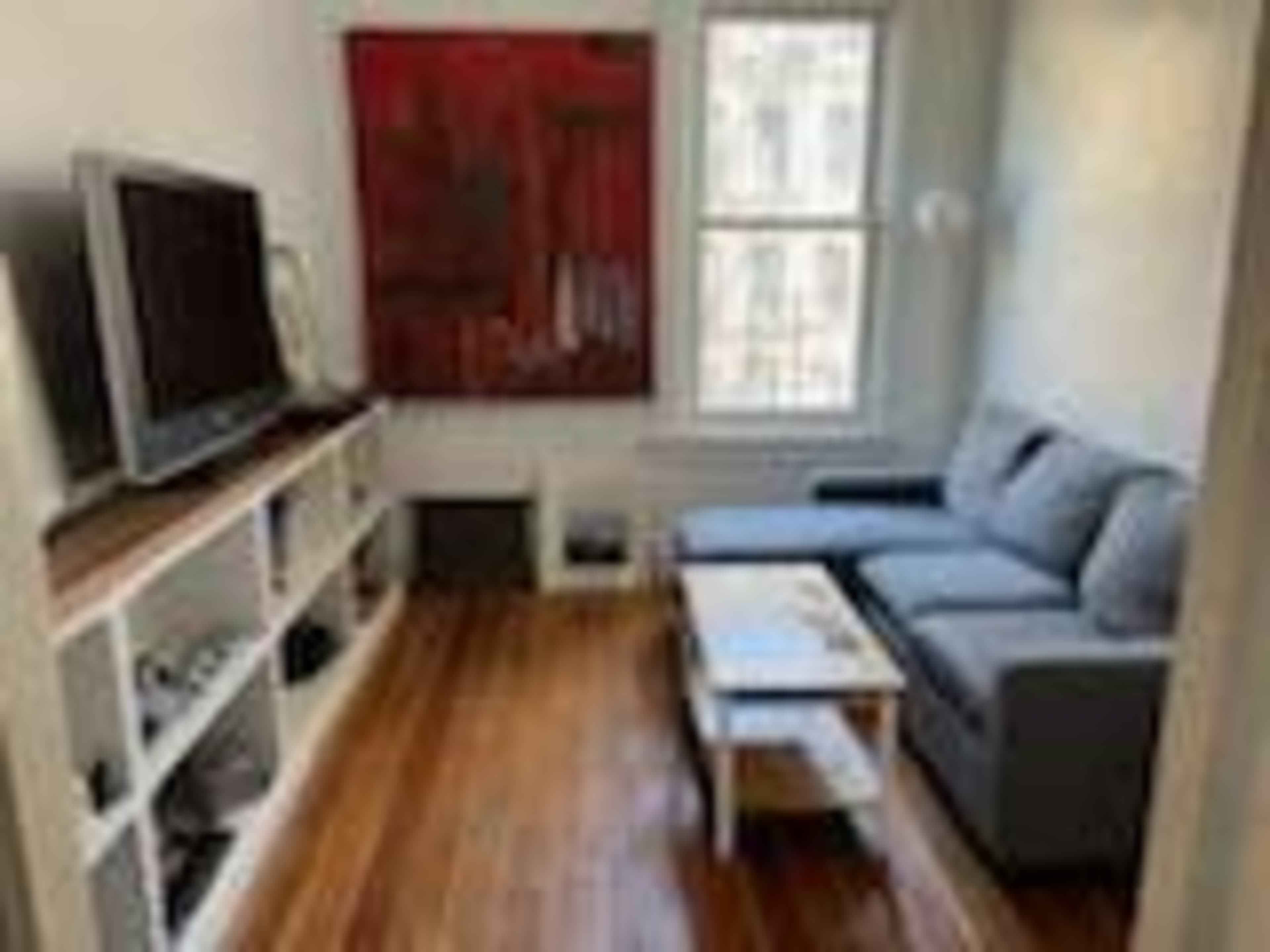 A living room features a gray sofa, a wooden coffee table, a television on a shelf, and a large red painting on the wall.
