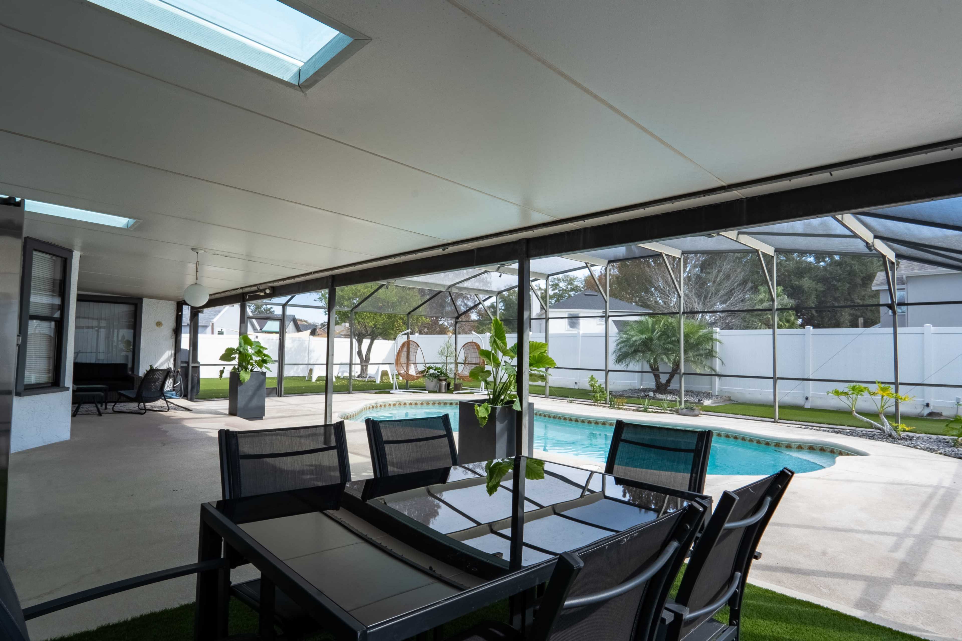 The image shows a screened-in patio with a dining table and chairs overlooking a swimming pool surrounded by greenery.