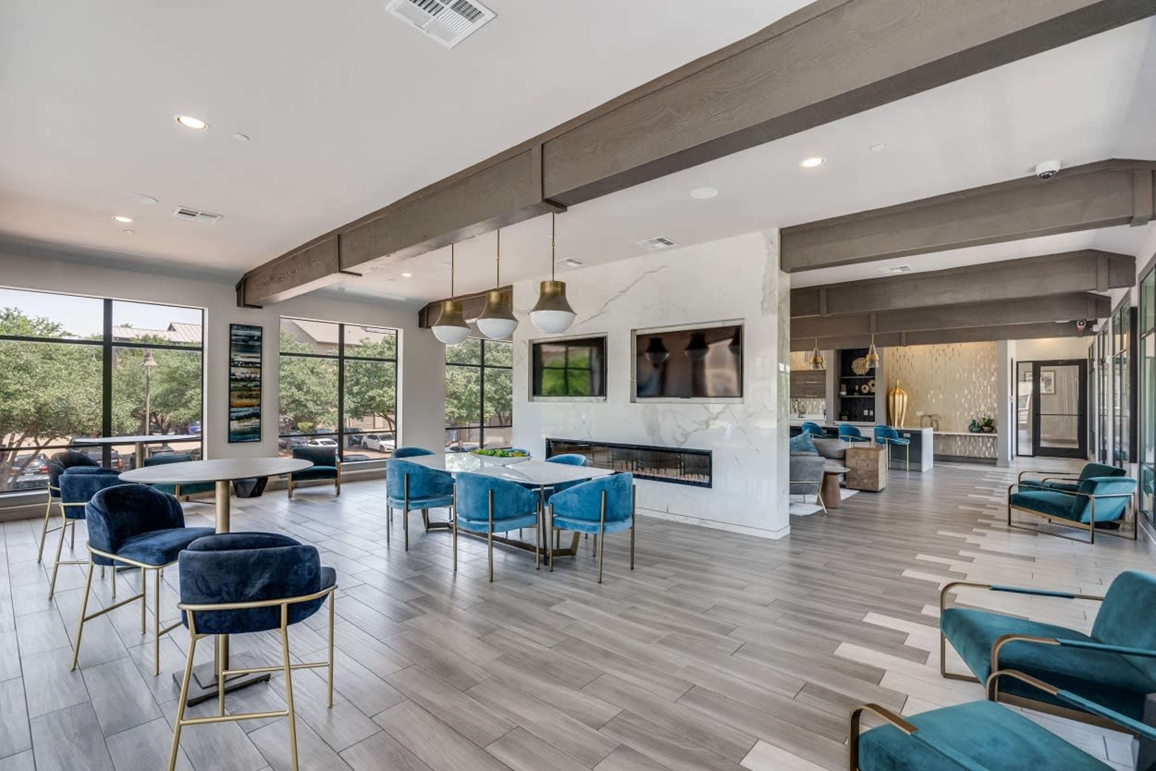 A modern common area with blue chairs, a fireplace, and large windows revealing trees outside.