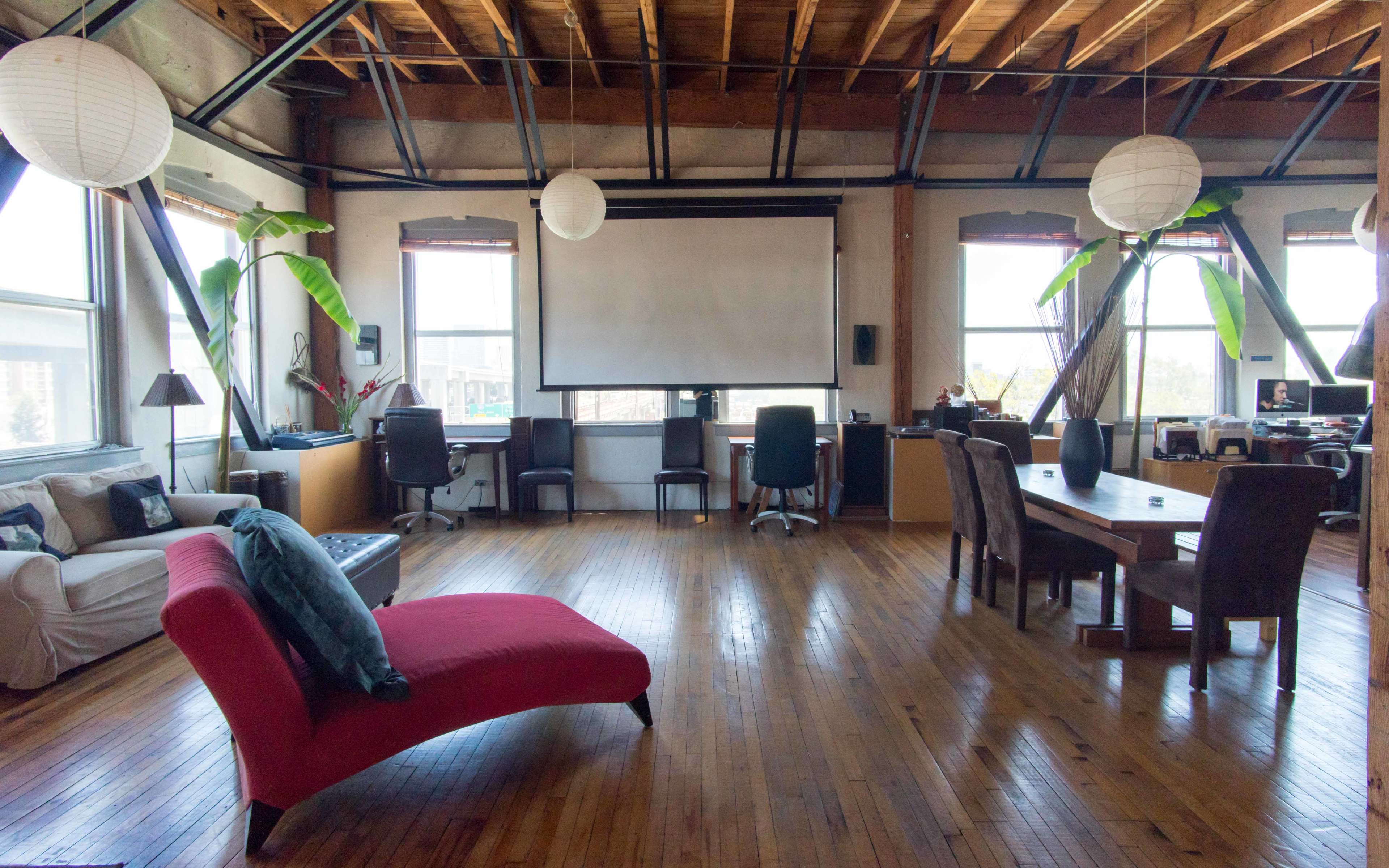 A spacious room with wooden flooring, large windows, a white projector screen, and several workstations surrounded by decorative paper lanterns and indoor plants.