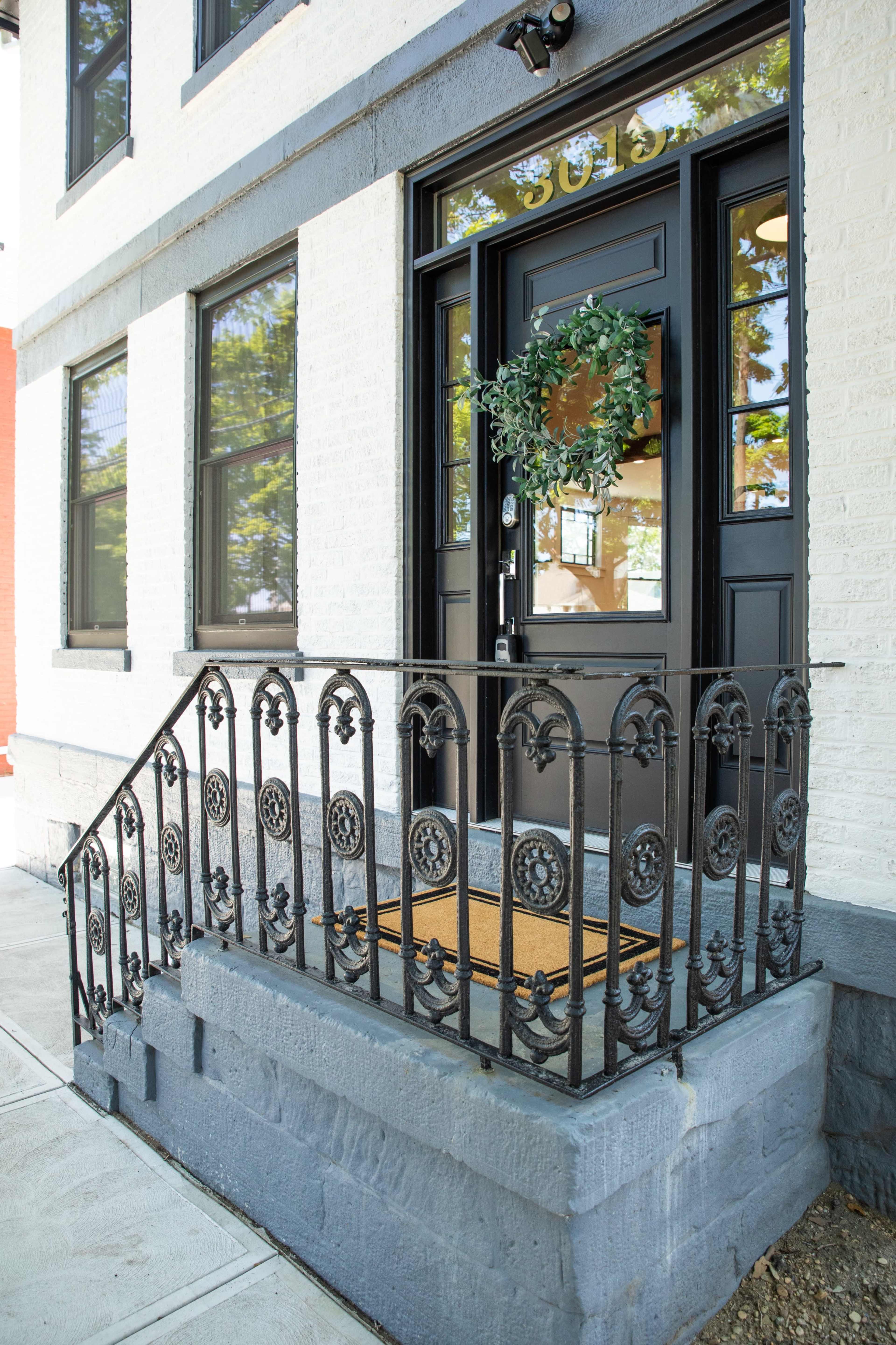 A black front door with a wreath, surrounded by a decorative iron railing and a small staircase leading to the entrance of a building.