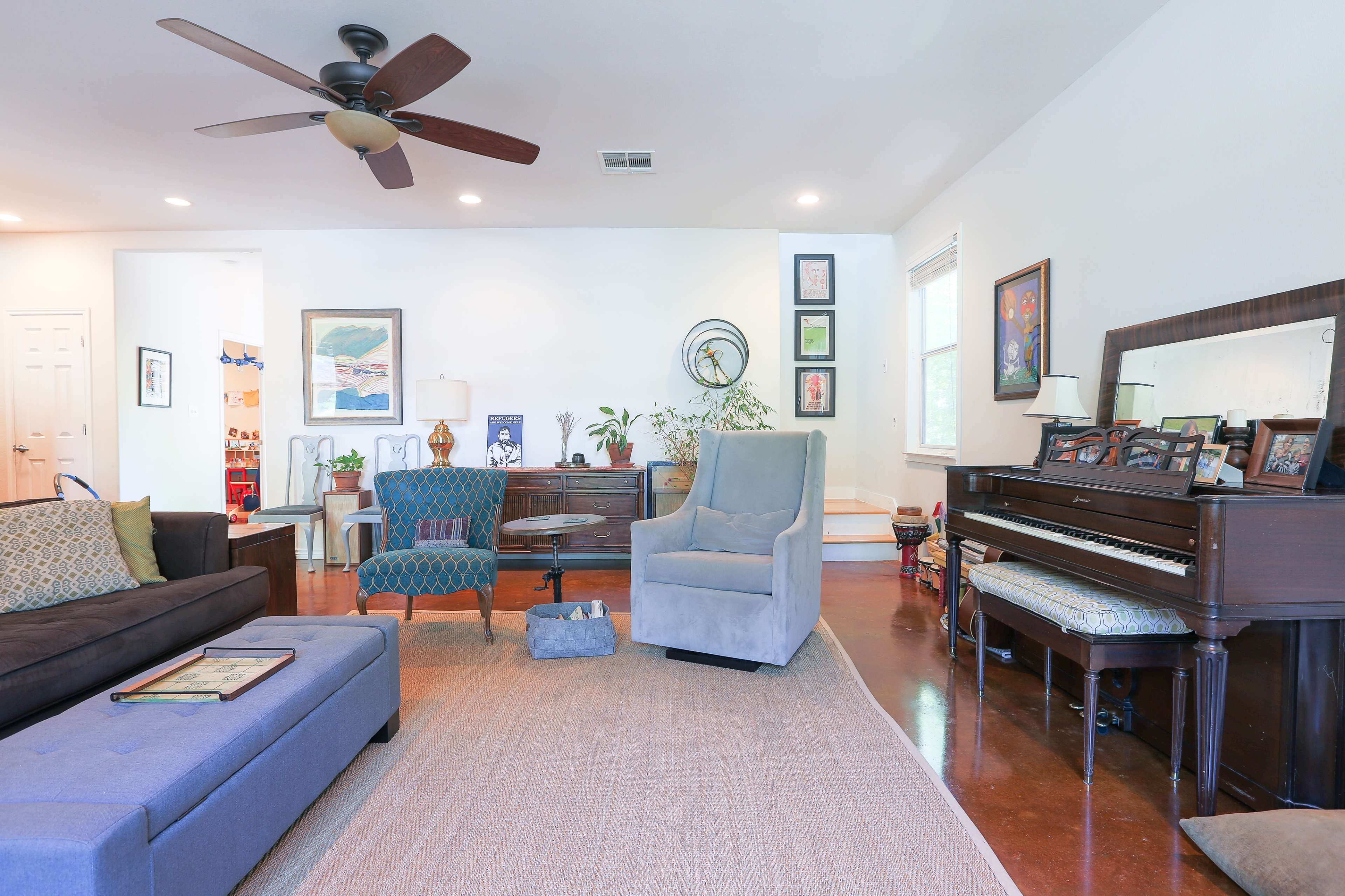 A spacious living room features a dark sofa, two upholstered chairs, a piano, and a large area rug.