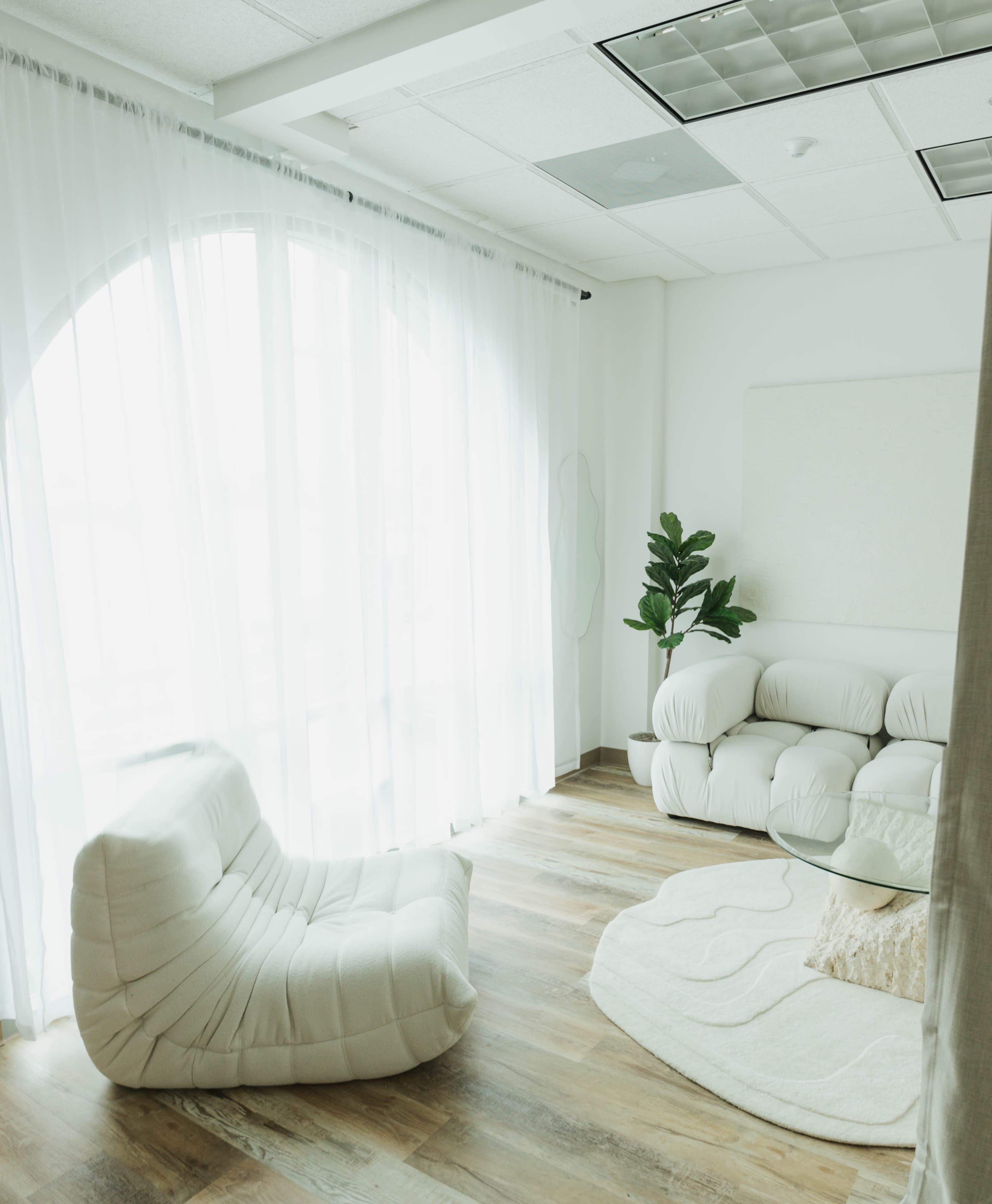 A minimalist, bright interior space with a curved seating area, a round coffee table, and a large window covered with sheer curtains.