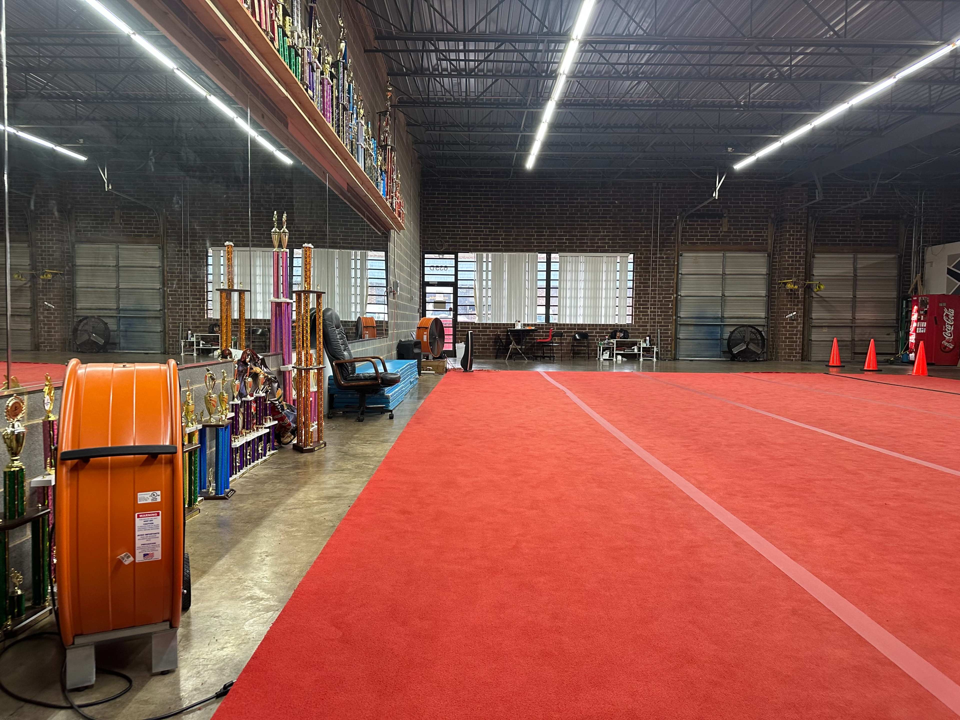 The image shows an indoor space with a red carpeted floor, decorated with various trophies along one wall, and large windows allowing daylight to enter.