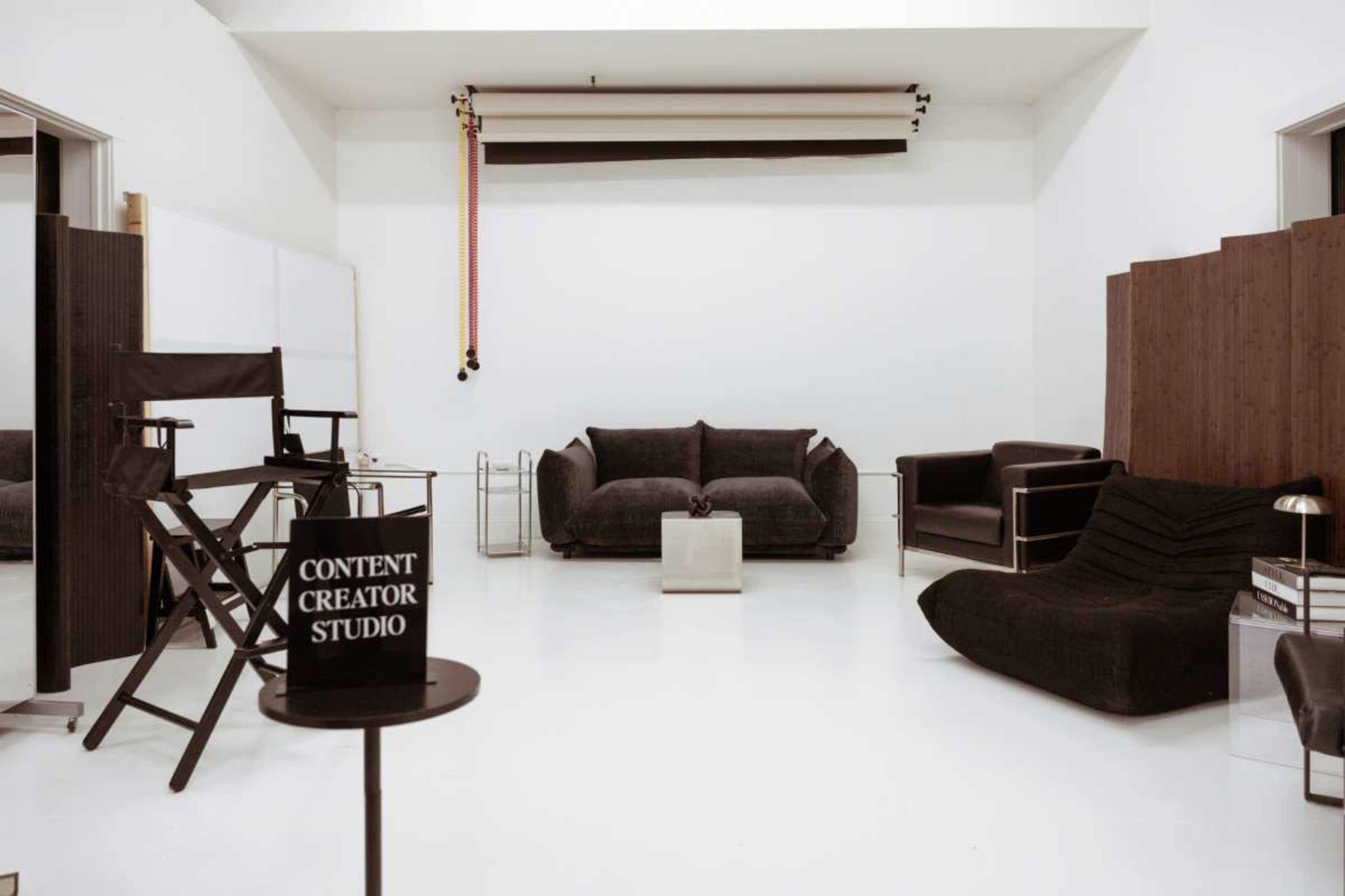 The image shows a minimalist studio space with white walls, two sofas, a director's chair, and a sign reading "CONTENT CREATOR STUDIO."