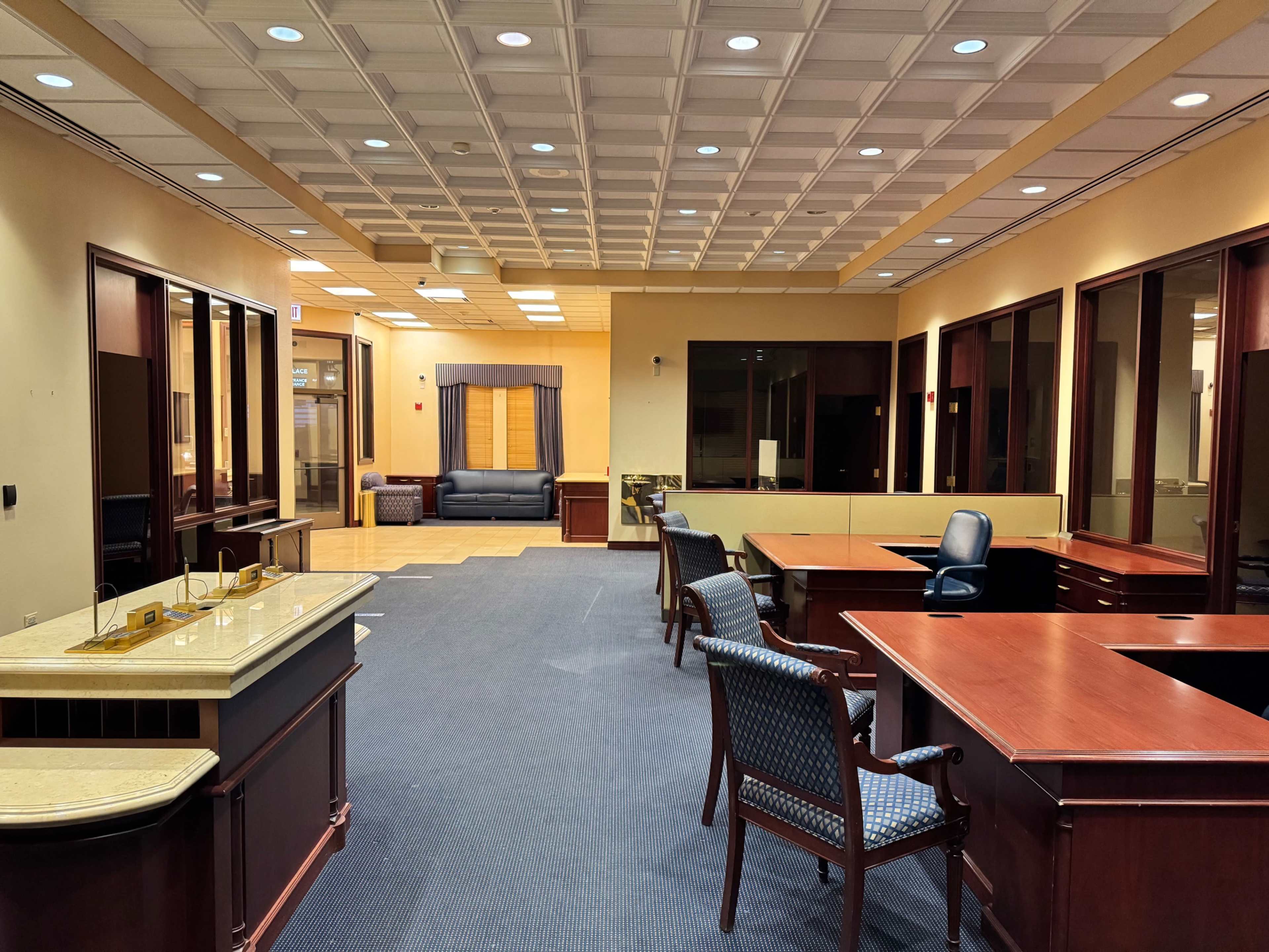 The image shows an interior office space featuring wooden furniture, including desks and chairs, with a carpeted floor and multiple windows allowing natural light.