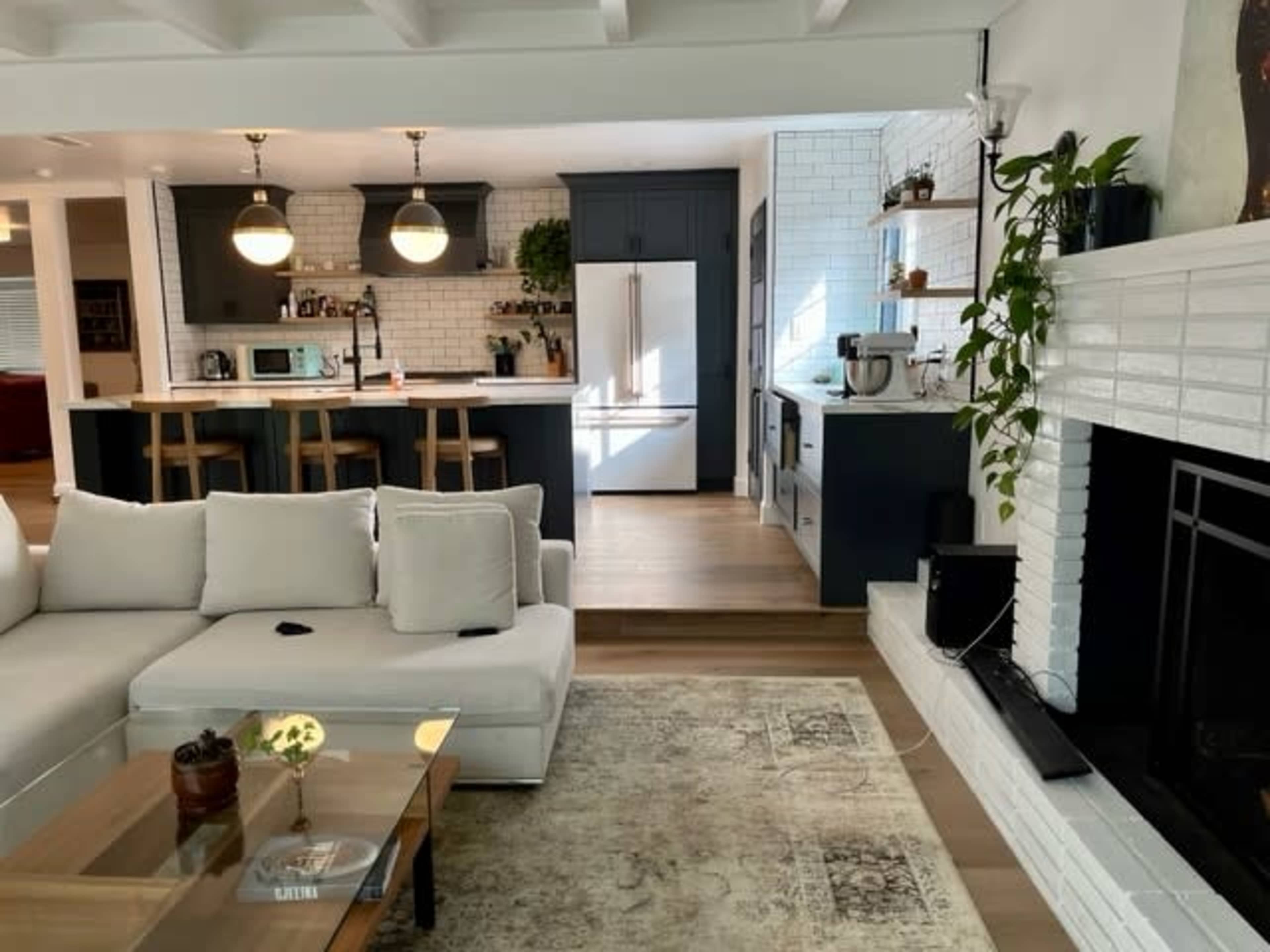 The image shows a modern living room featuring a white sectional sofa, a wooden coffee table, and a kitchen area with dark cabinetry and white tiles in the background.