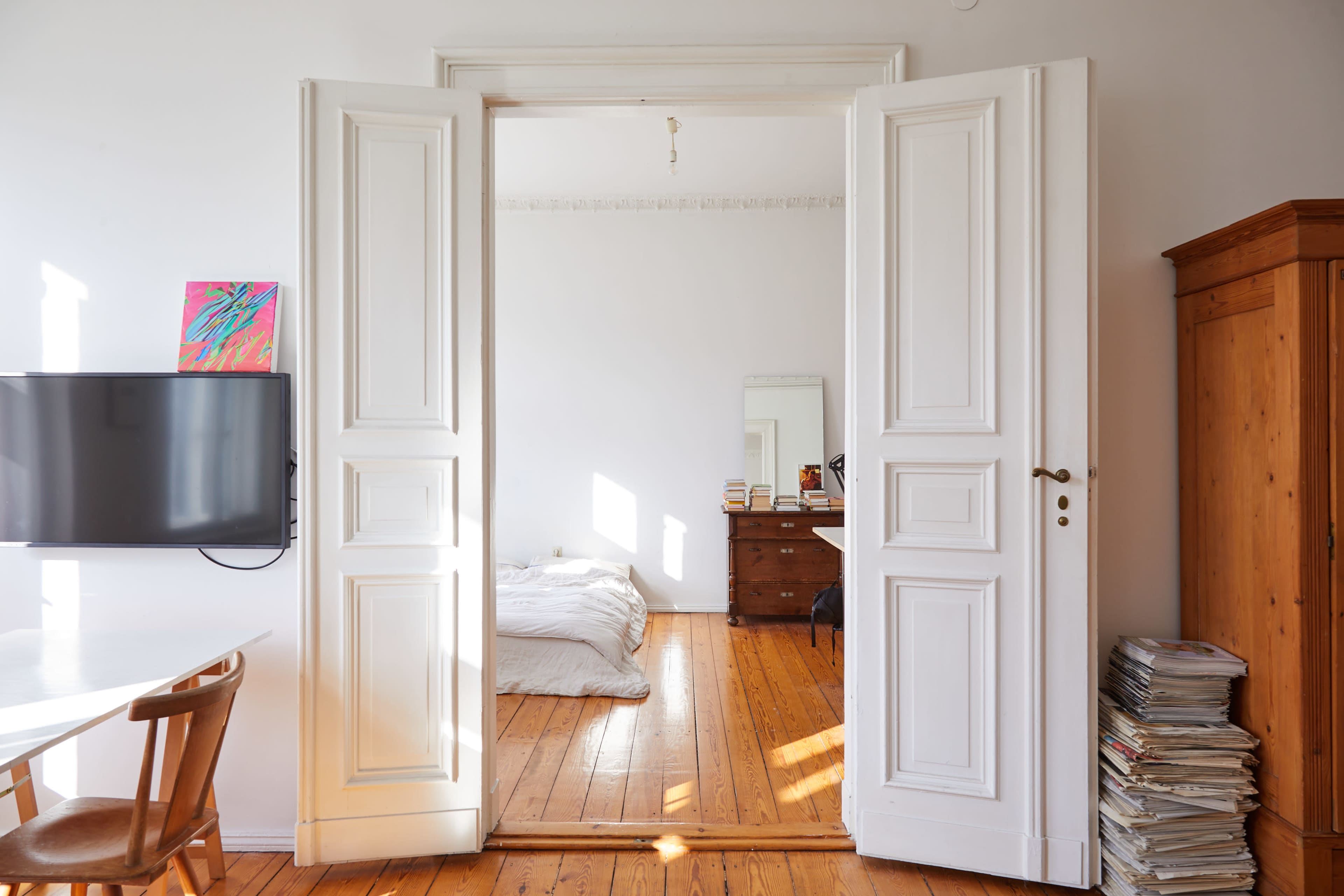 A pair of white double doors opens to a bedroom featuring a wooden dresser and a bed on a wooden floor.