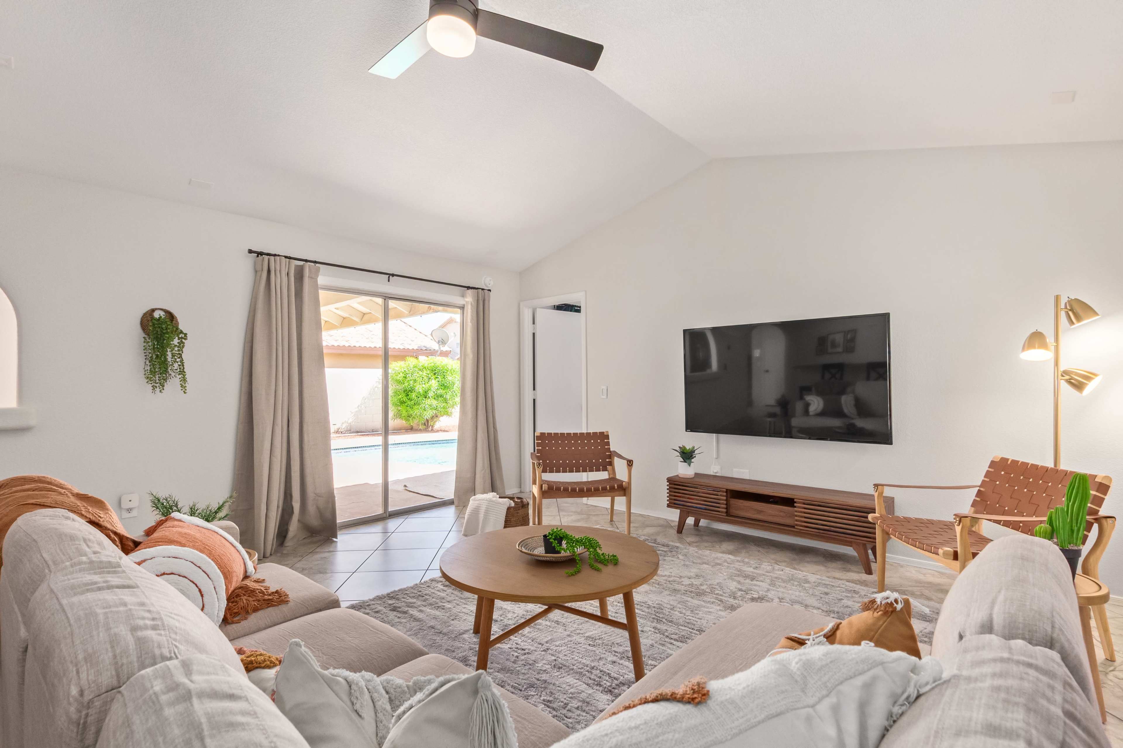 The living room features a sectional sofa, a round coffee table, and a large TV, with sliding glass doors leading to a pool area.