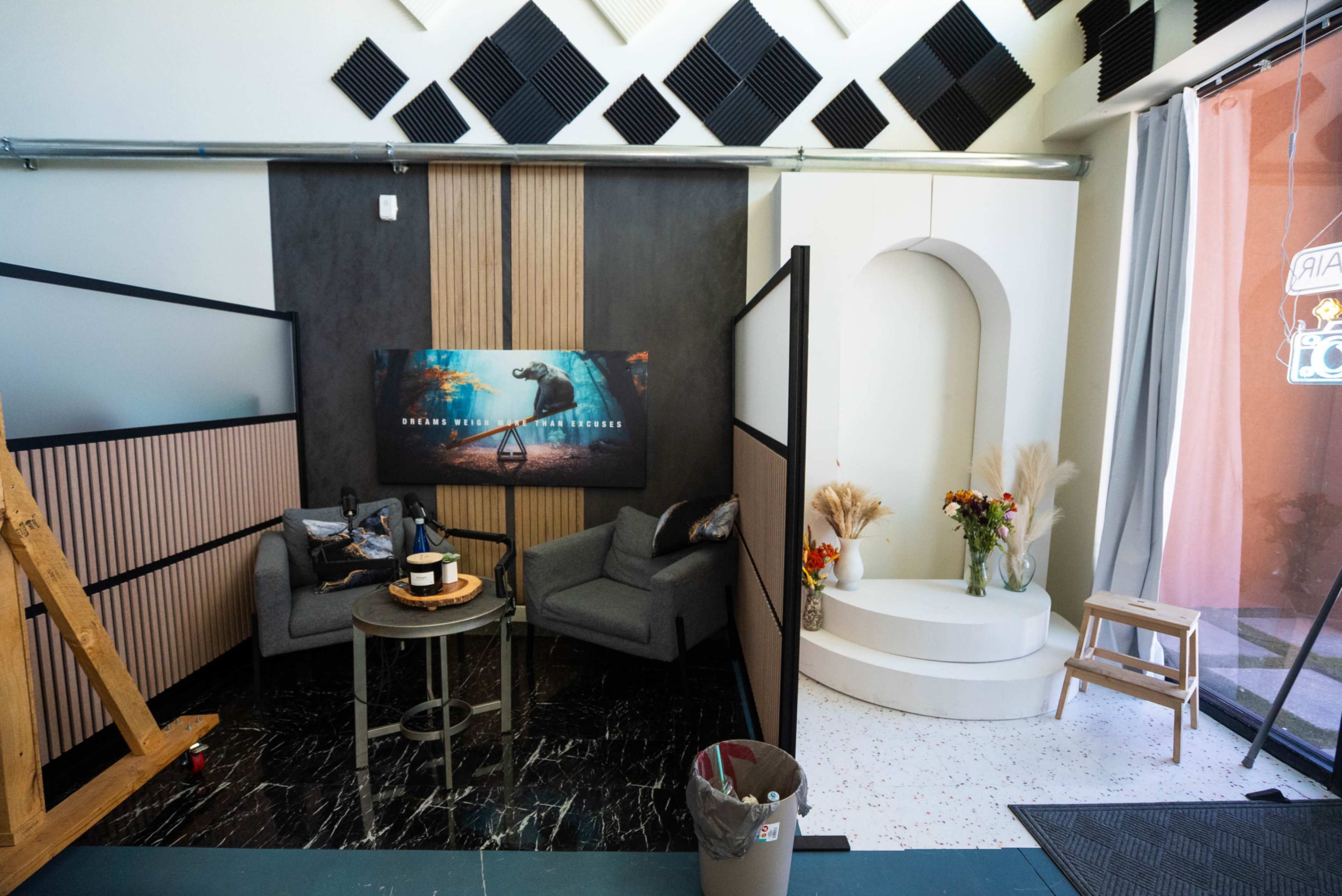 The image depicts a modern interior space featuring two grey armchairs, a small coffee table, a decorative backdrop with a large poster, and a curved white platform in the corner.