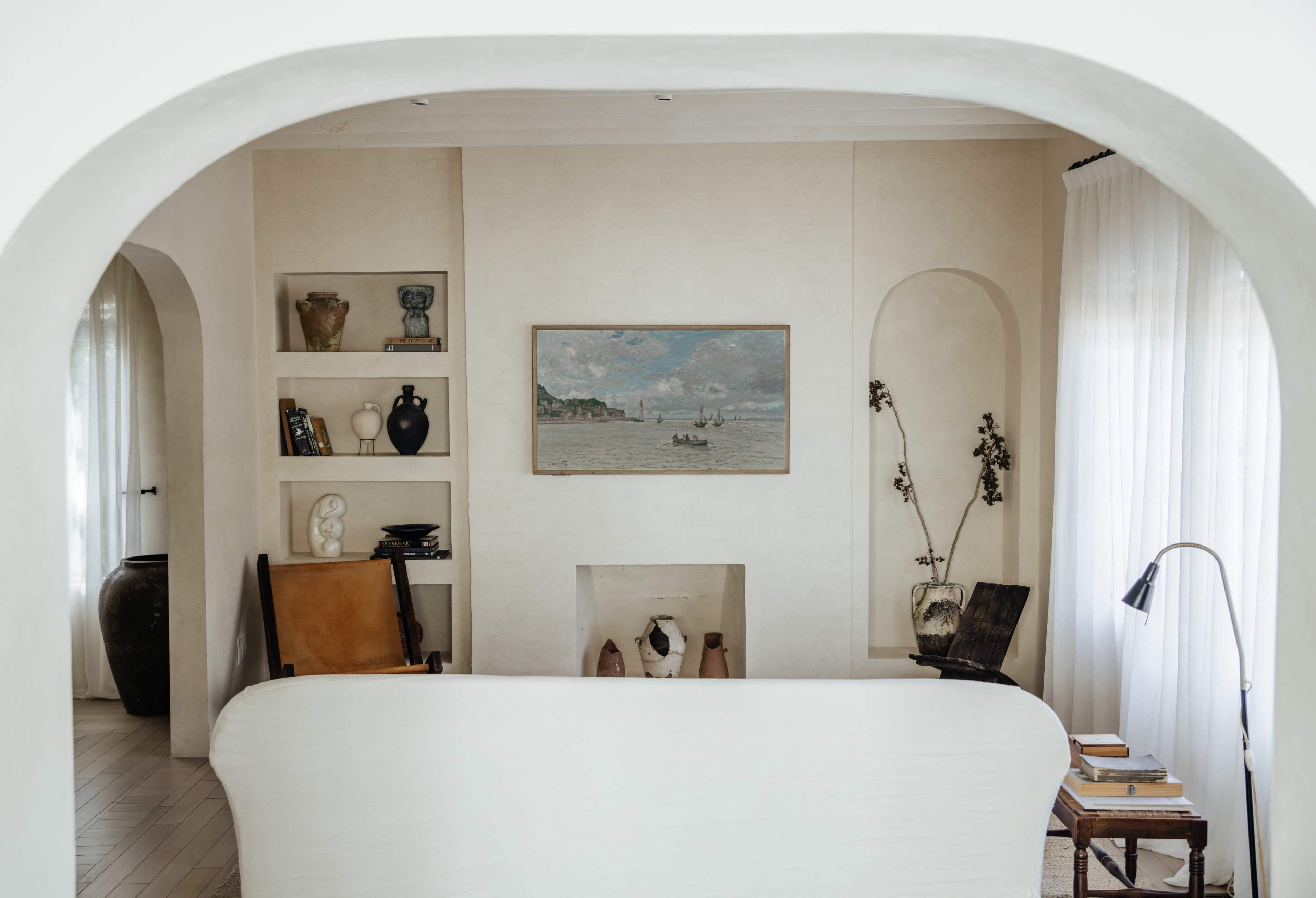 A light and airy living room features a large painting, shelves with decorative items, and a variety of seating arrangements.