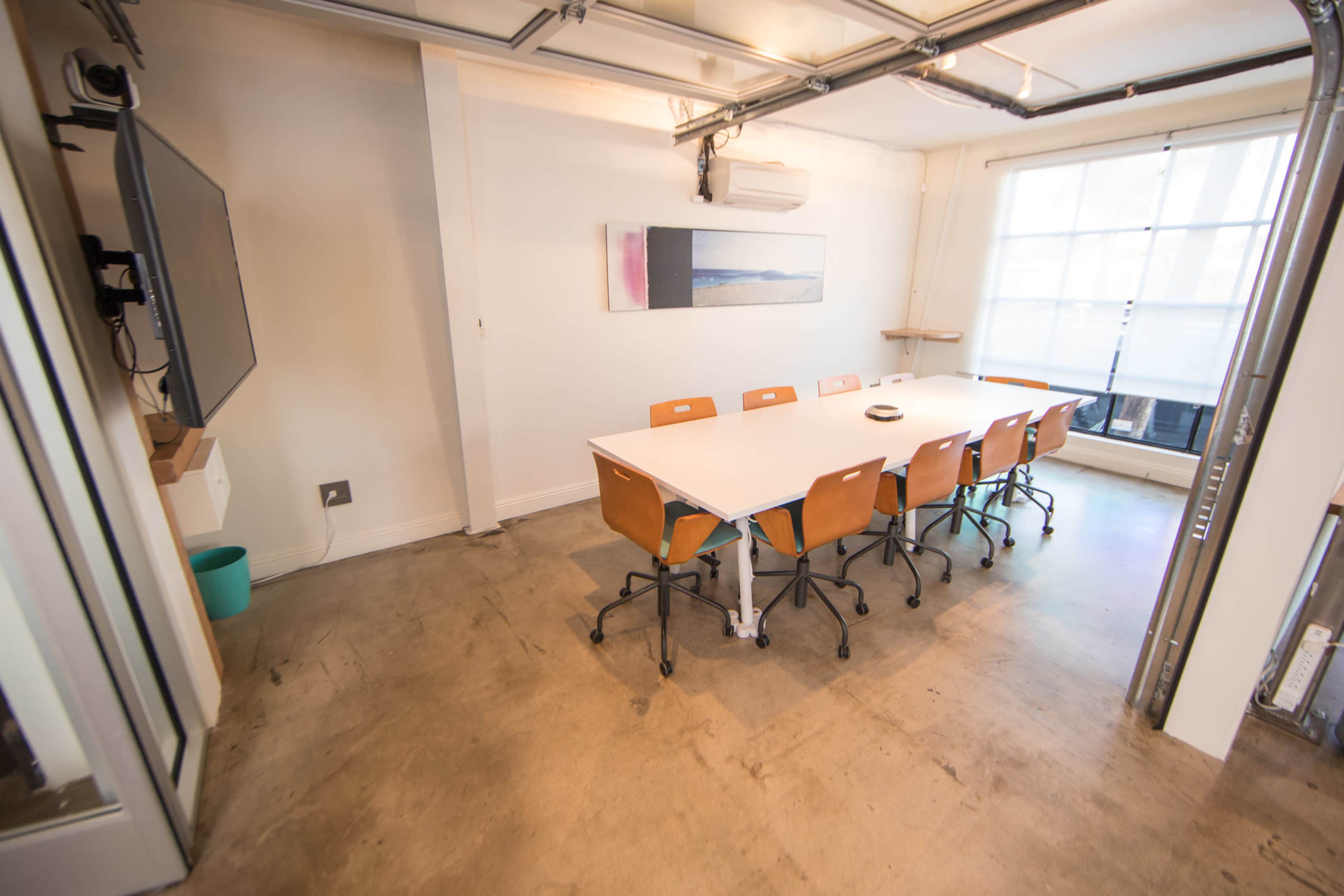 A minimalistic meeting room features a large white table surrounded by orange chairs, with a television mounted on the wall and a window letting in natural light.