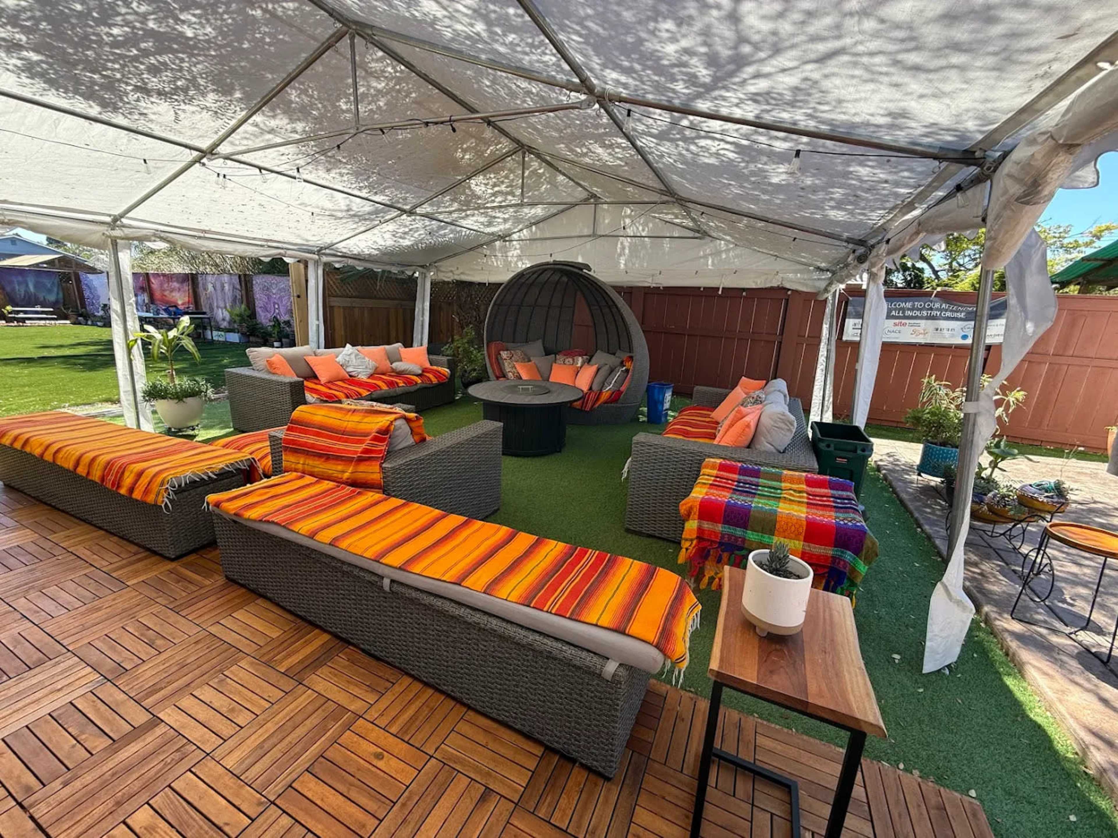 A tented outdoor lounge area features several couches with vibrant striped blankets, a round seating pod, and potted plants on a wooden floor.