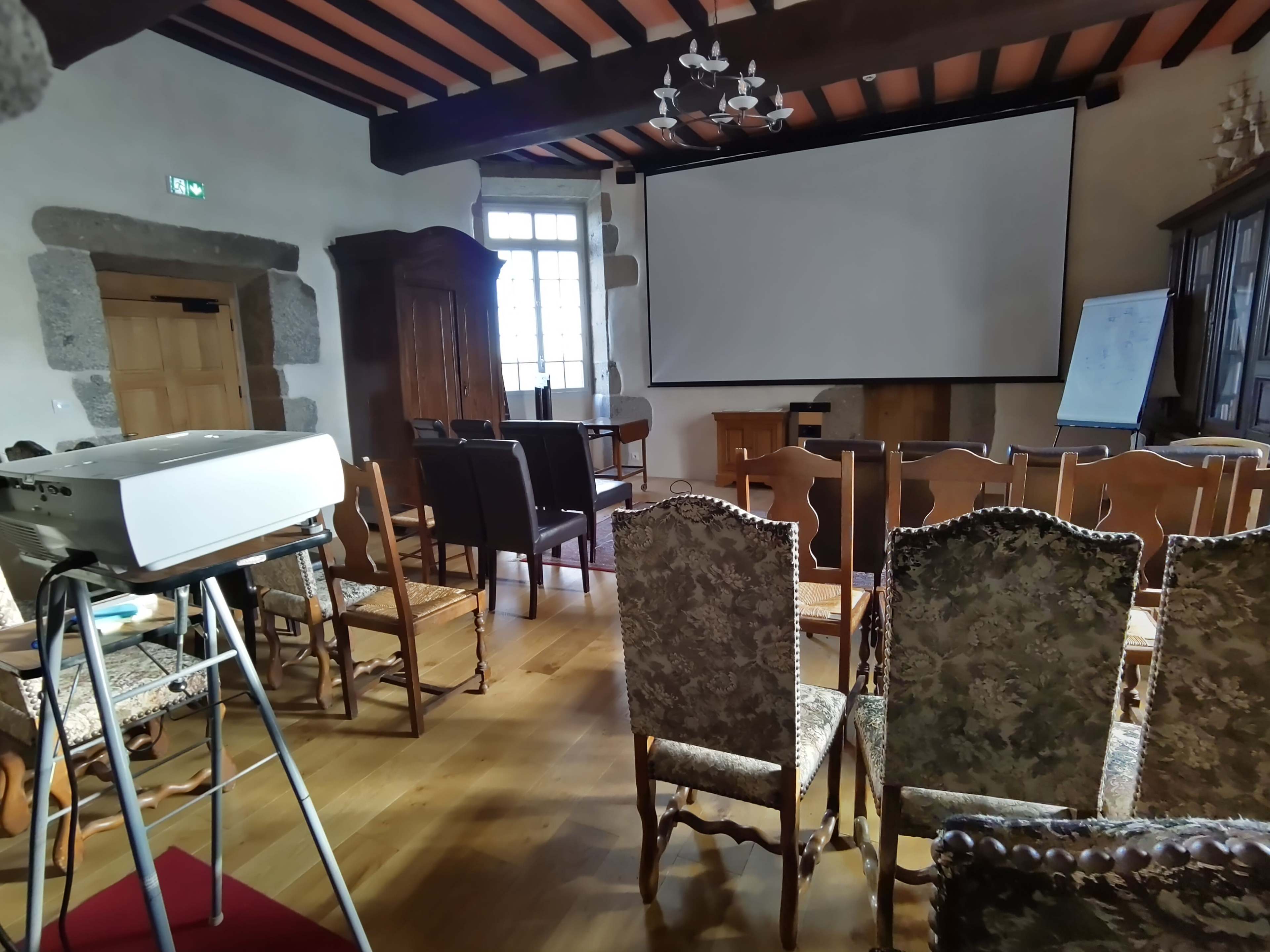 The image shows a classroom or meeting room with chairs arranged in rows, a projector on a stand, and a large screen in front.