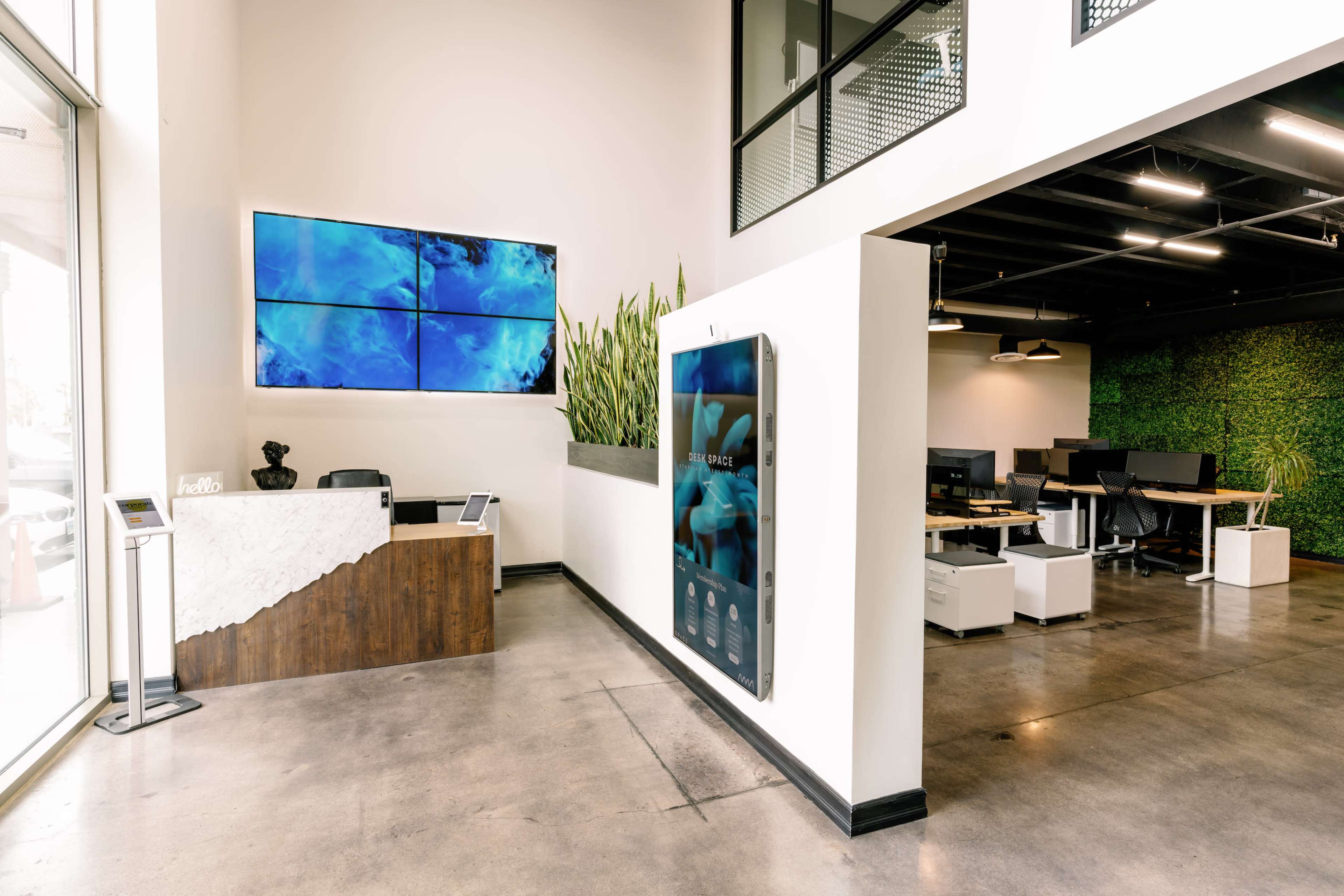 A modern office reception area with a wooden front desk, digital displays on the walls, and lush green plants in the background.