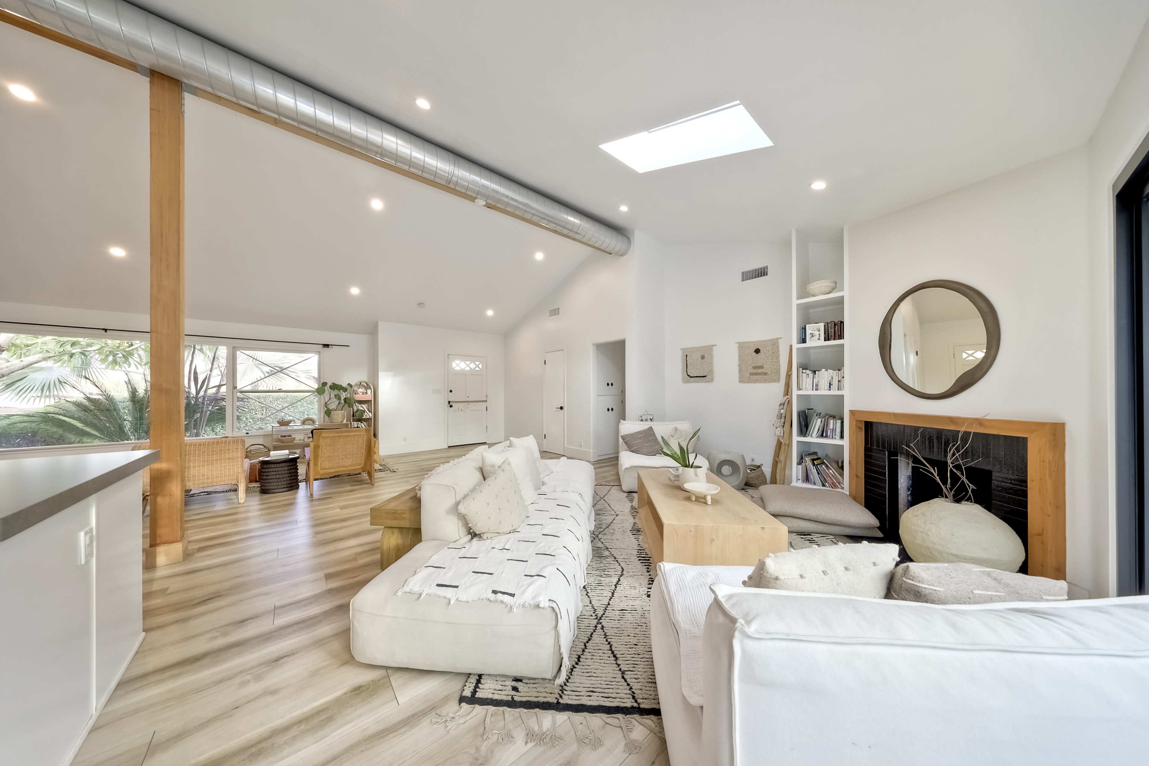 The image shows a modern, open-concept living room with a light-colored sofa, a wooden coffee table, and large windows allowing natural light to enter.