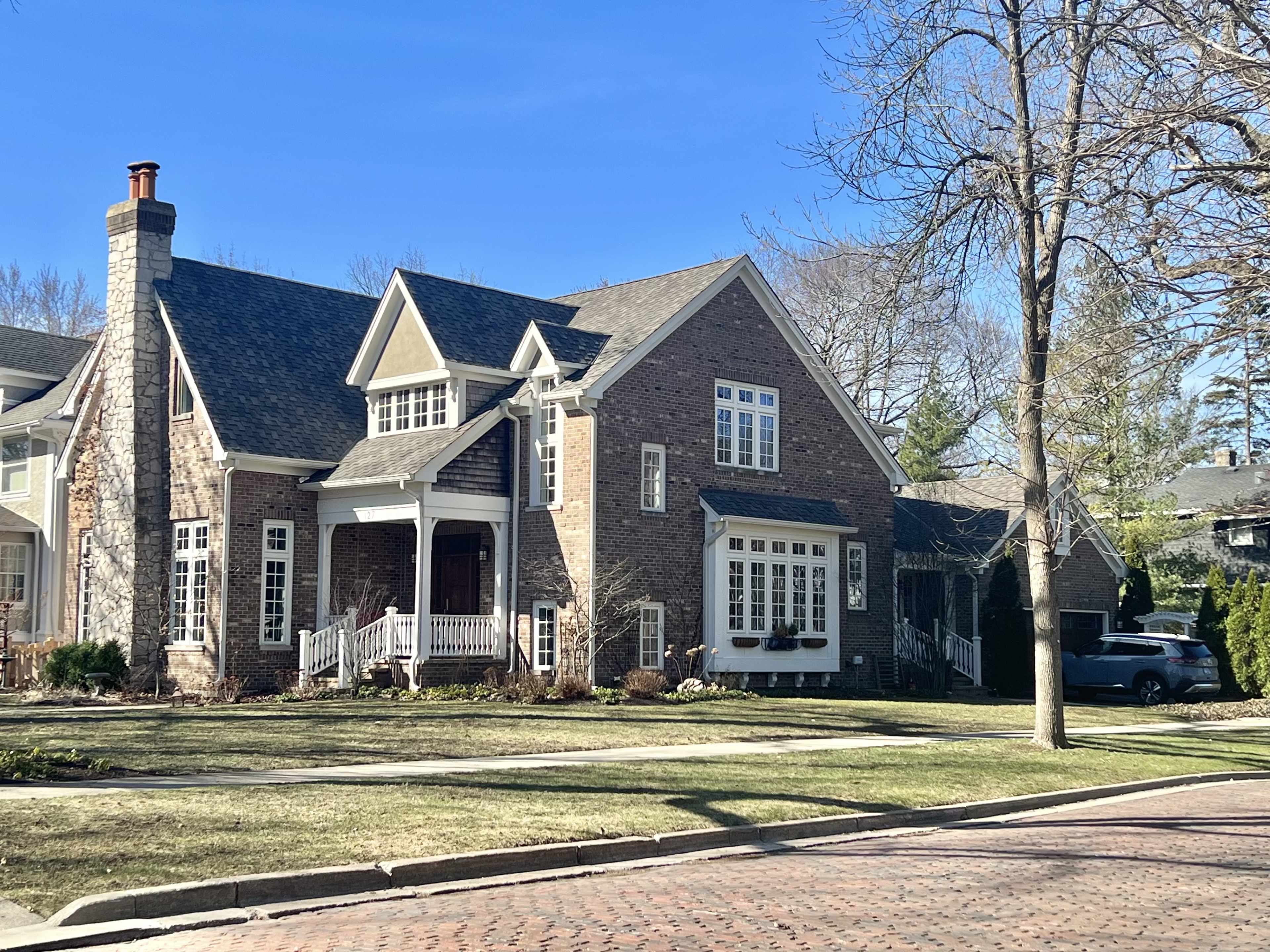 A large, two-story brick house with a stone façade and multiple gables is situated on a well-maintained street.