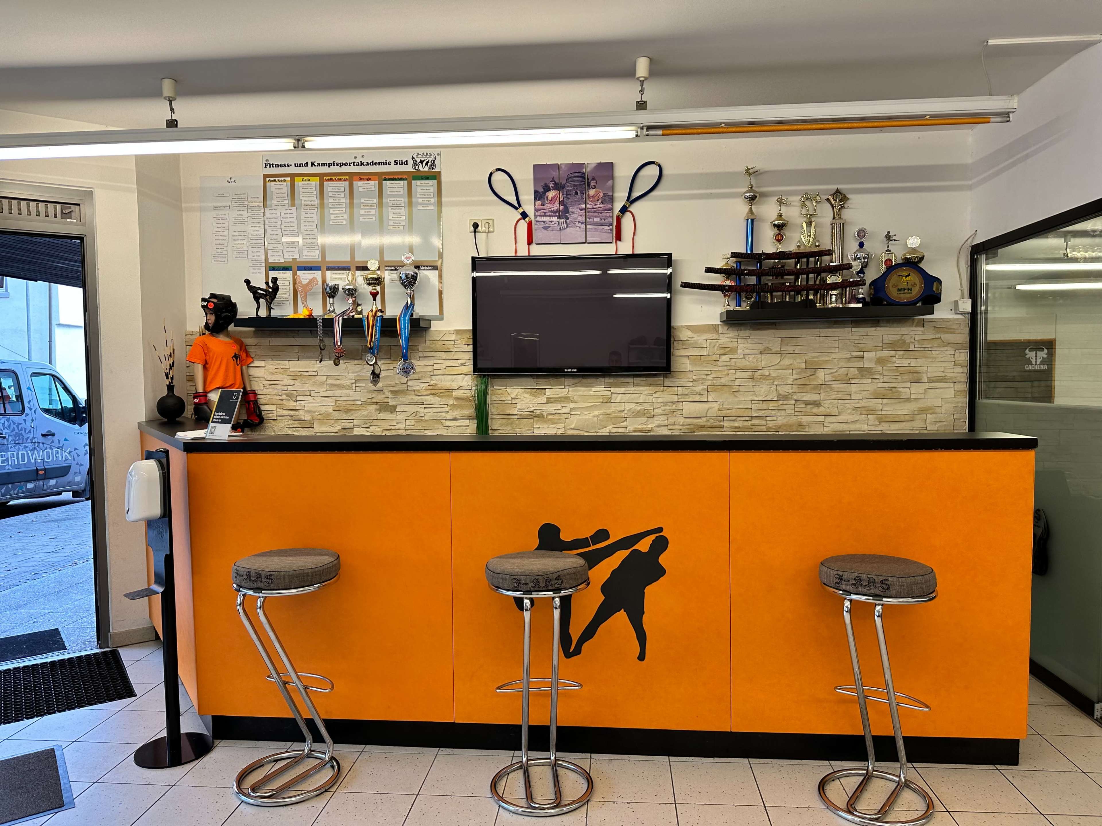The image shows a reception area with an orange counter, stools, a television, and various trophies displayed on a stone wall background.