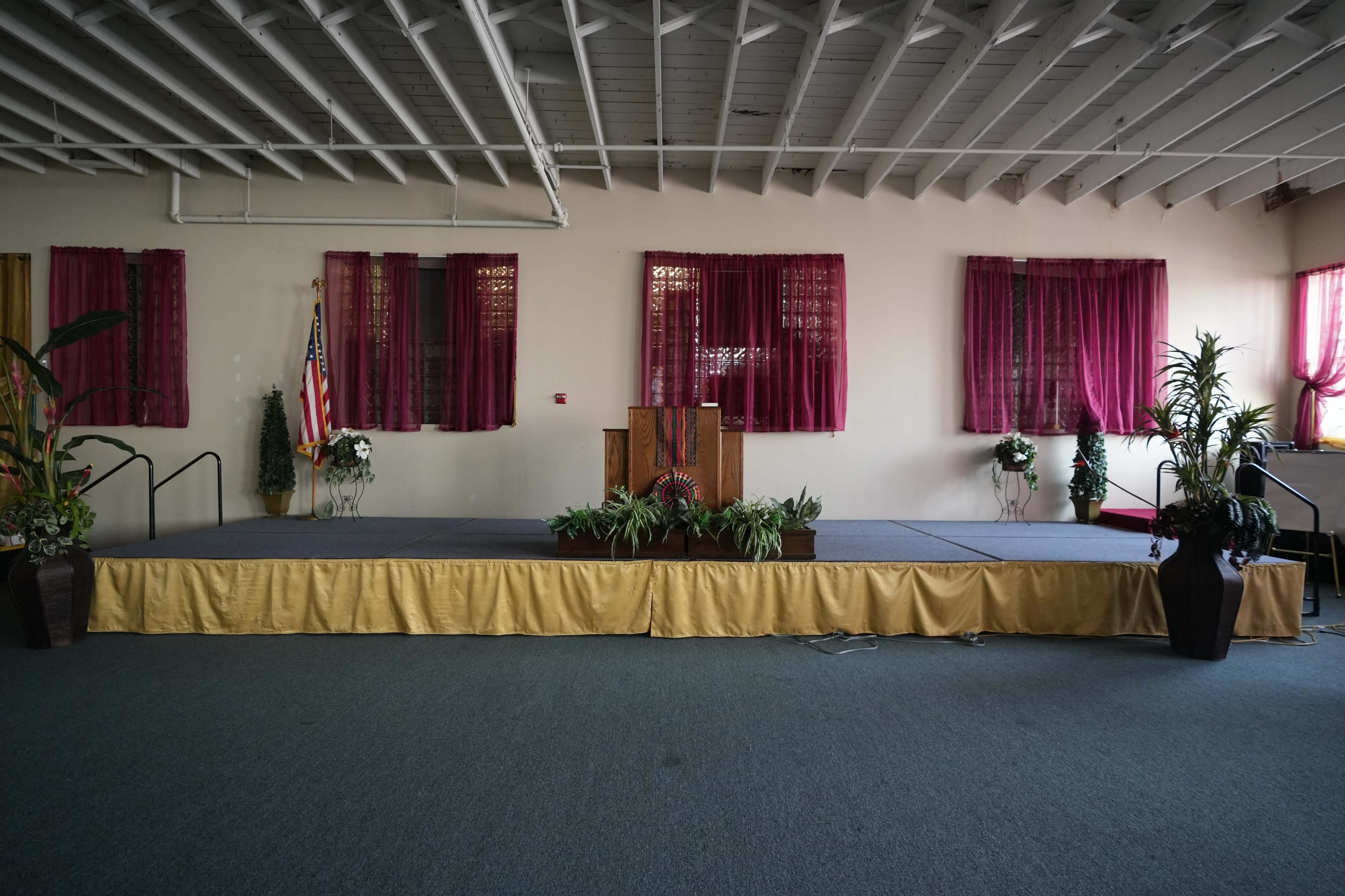 The image shows a stage with a podium at the front of a spacious room, decorated with potted plants and curtains.