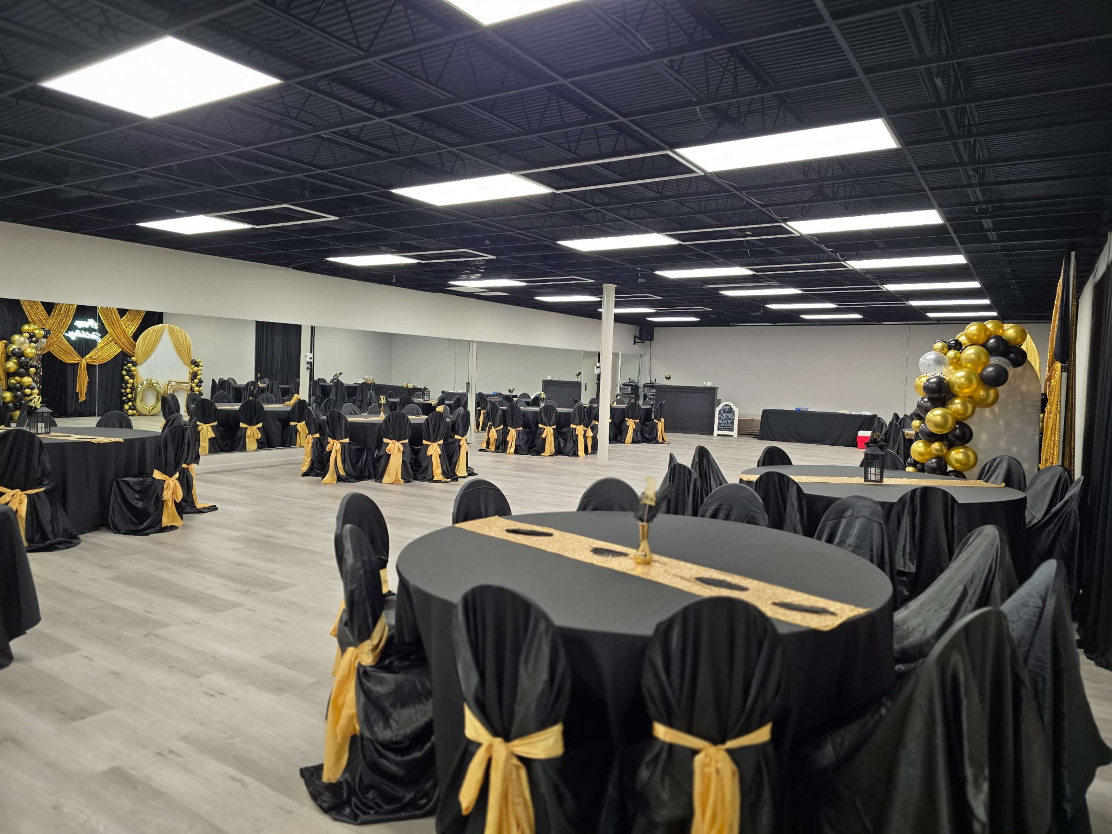 The image shows a large event space set with round tables covered in black tablecloths and gold accents, surrounded by chairs draped with black fabric and gold ribbons.