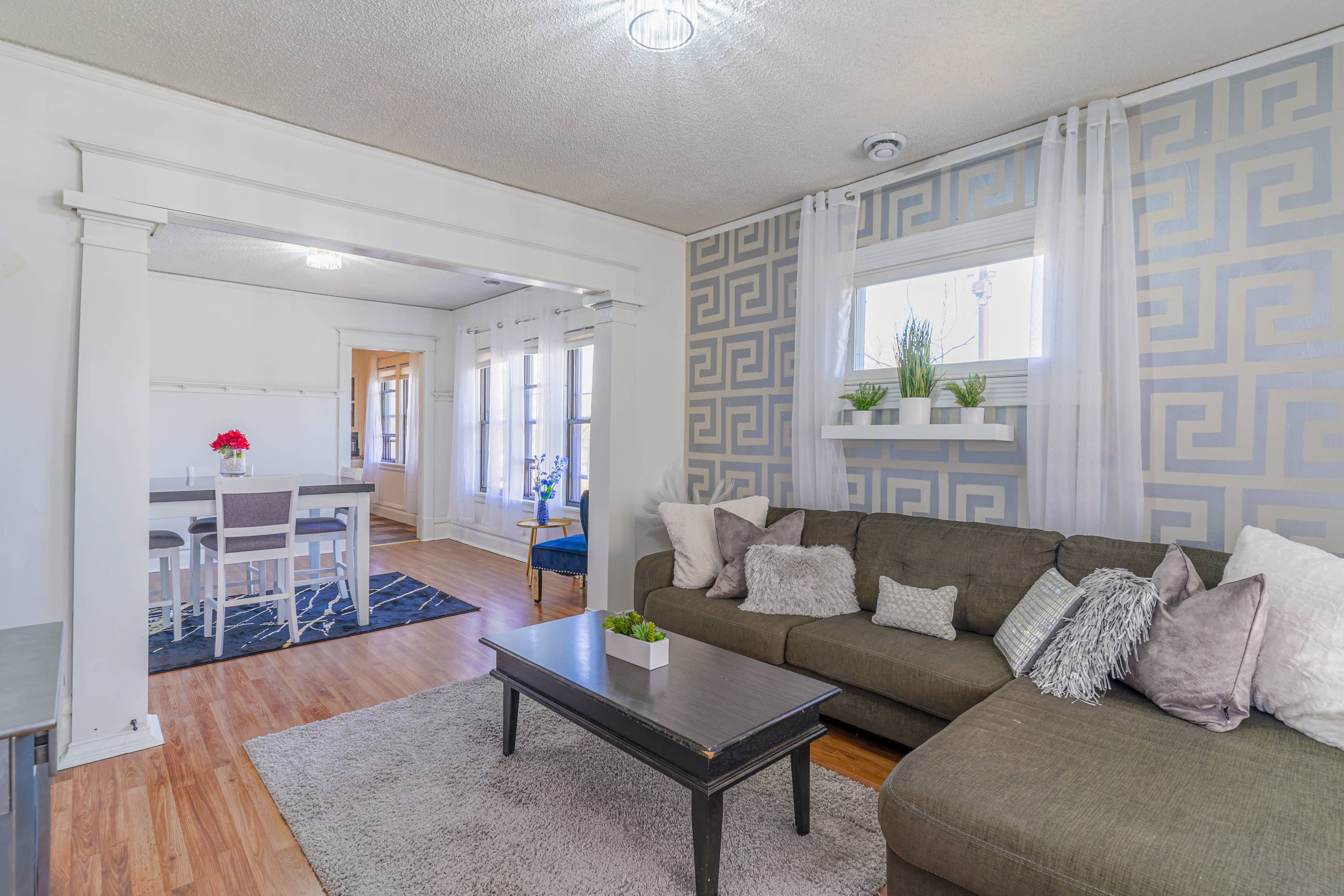 The image shows a living room with a sectional sofa, a coffee table, and decorative throw pillows, leading into a dining area with a white table and chairs.