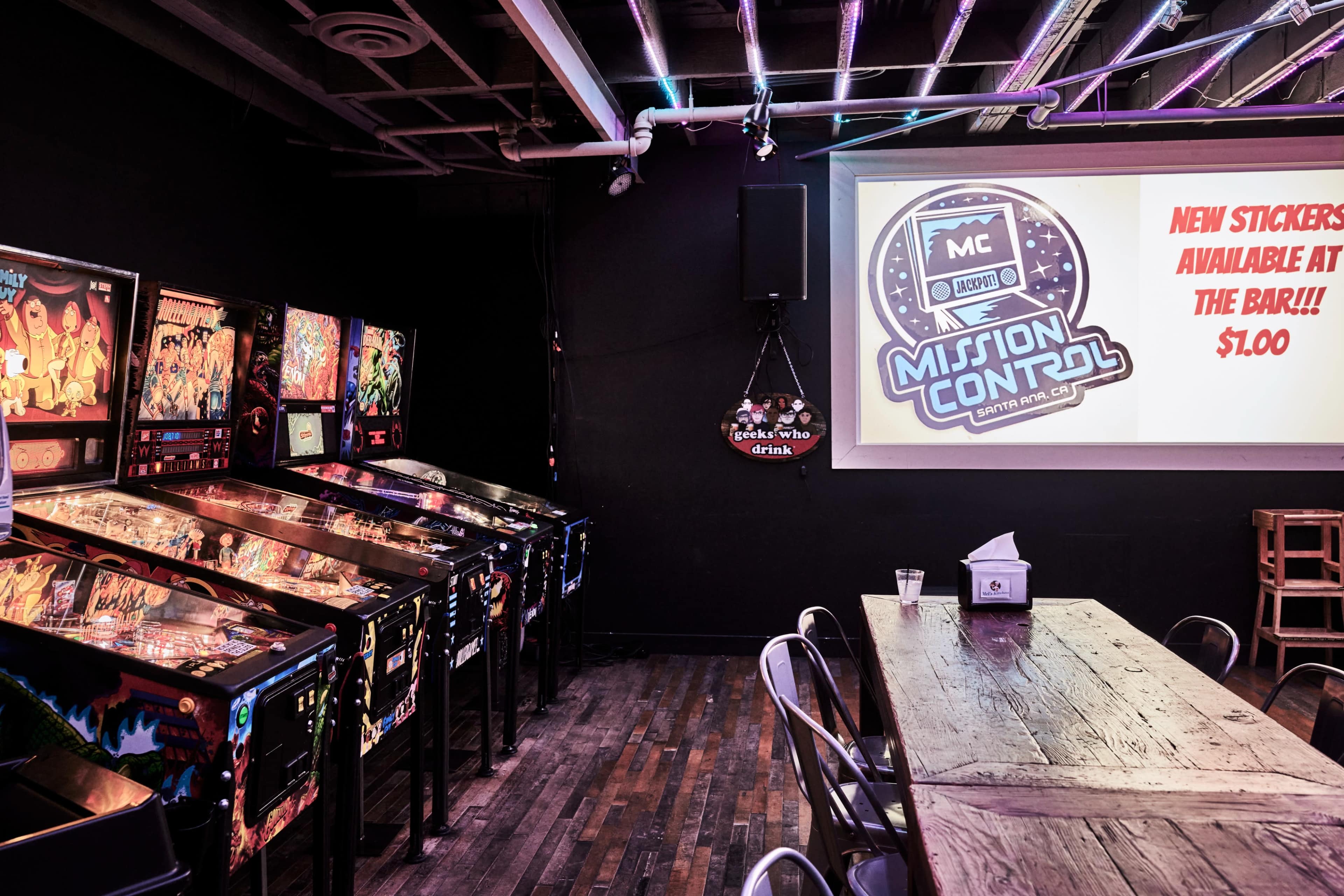 A dimly lit arcade room featuring several pinball machines along one wall and a large screen displaying a sign about stickers for sale at the bar.