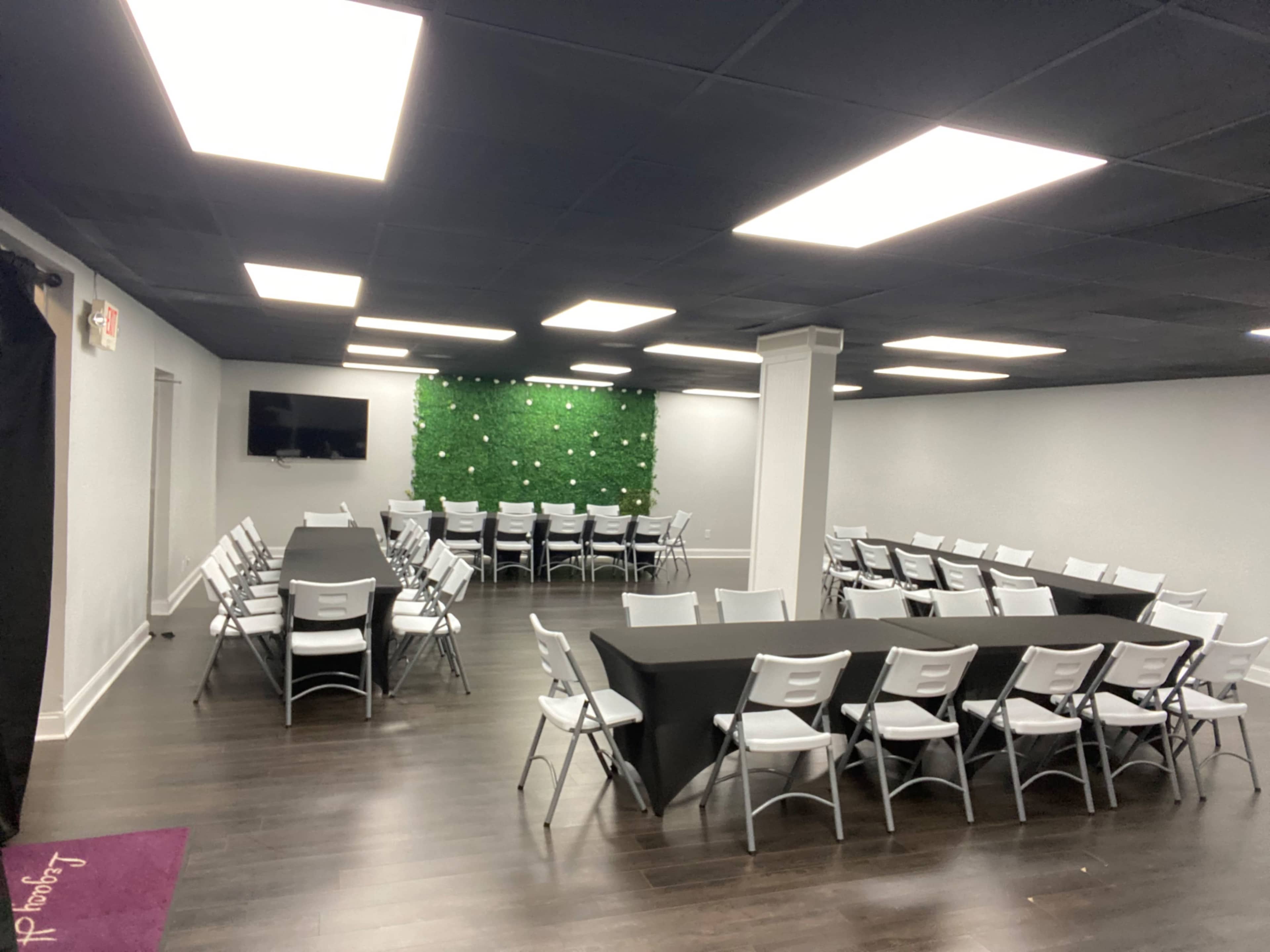 A large, well-lit conference room arranged with several rows of white chairs and tables, along with a decorative green wall at one end.
