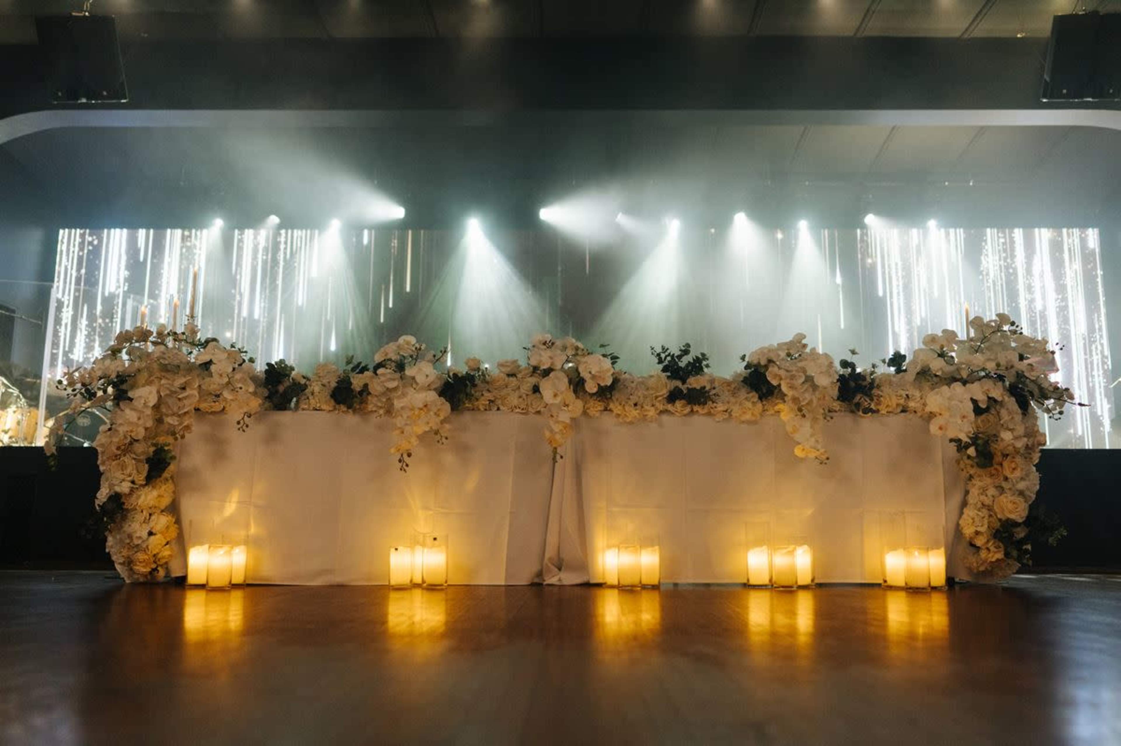 A decorated table adorned with white flowers and candles is illuminated by soft lighting in a dimly lit venue.