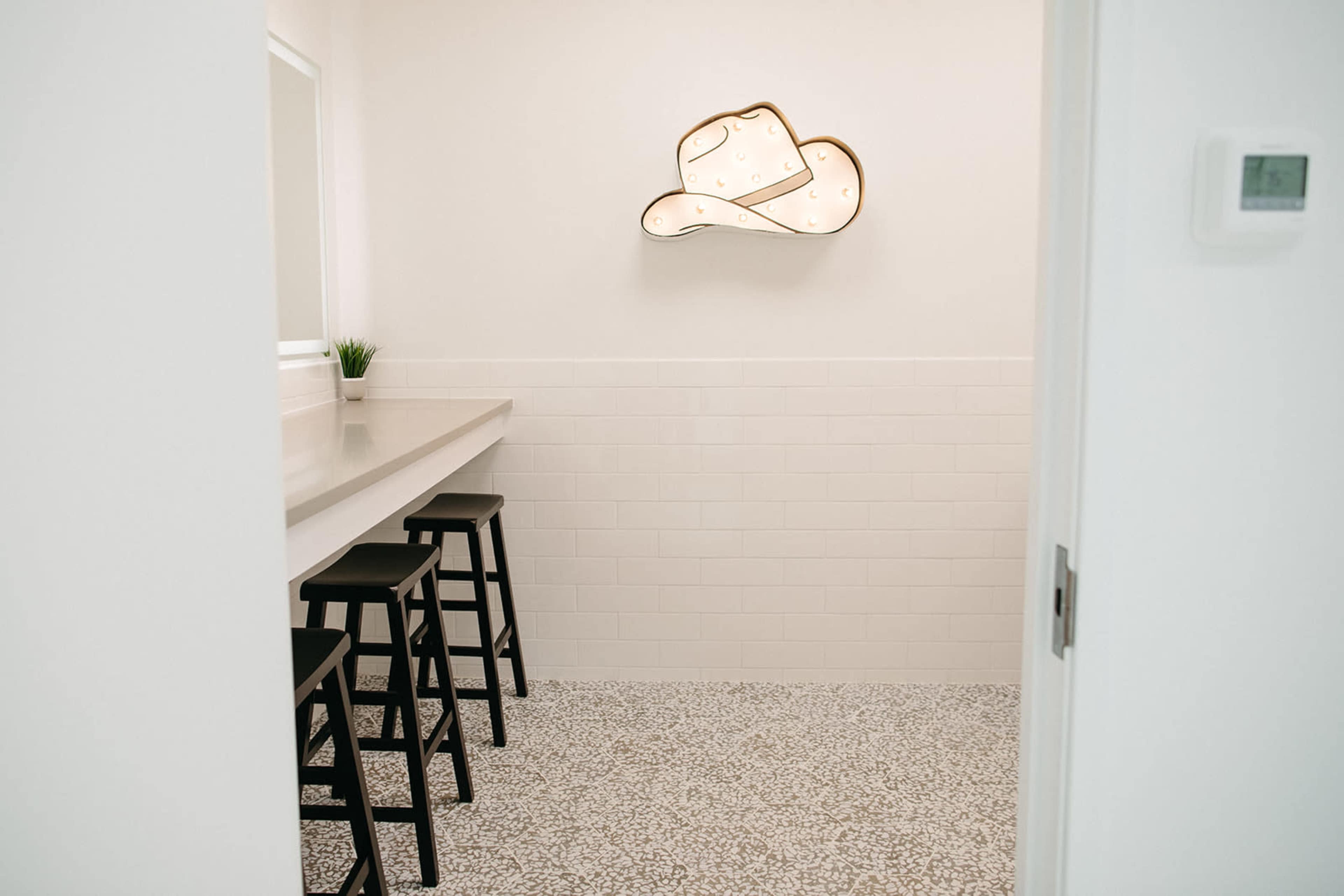 A small room with a lighted cowboy hat decoration on the wall, a long counter with four black stools, and a white tiled backsplash.