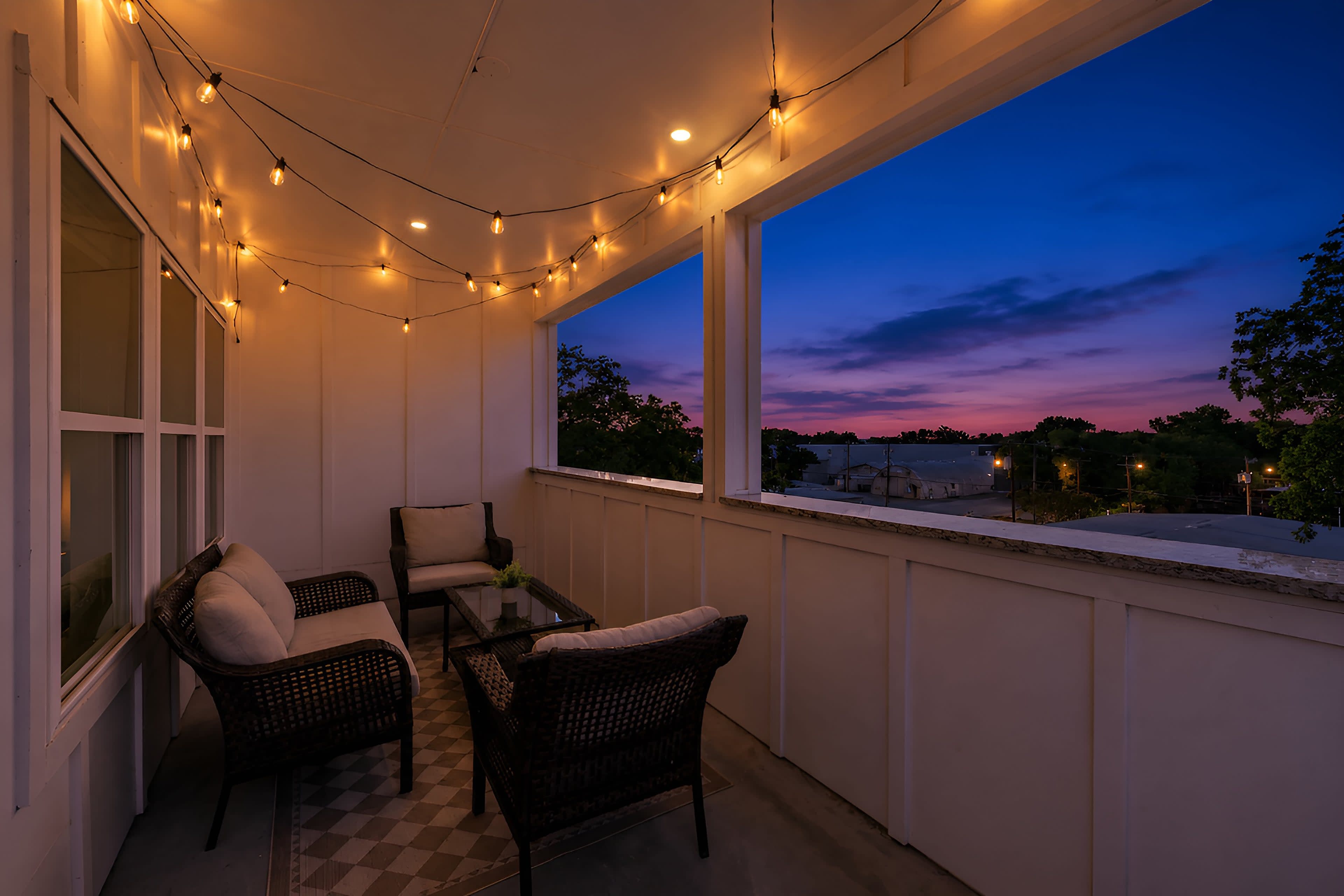 A cozy outdoor balcony features string lights illuminating two chairs and a small table, with a scenic sunset view in the background.