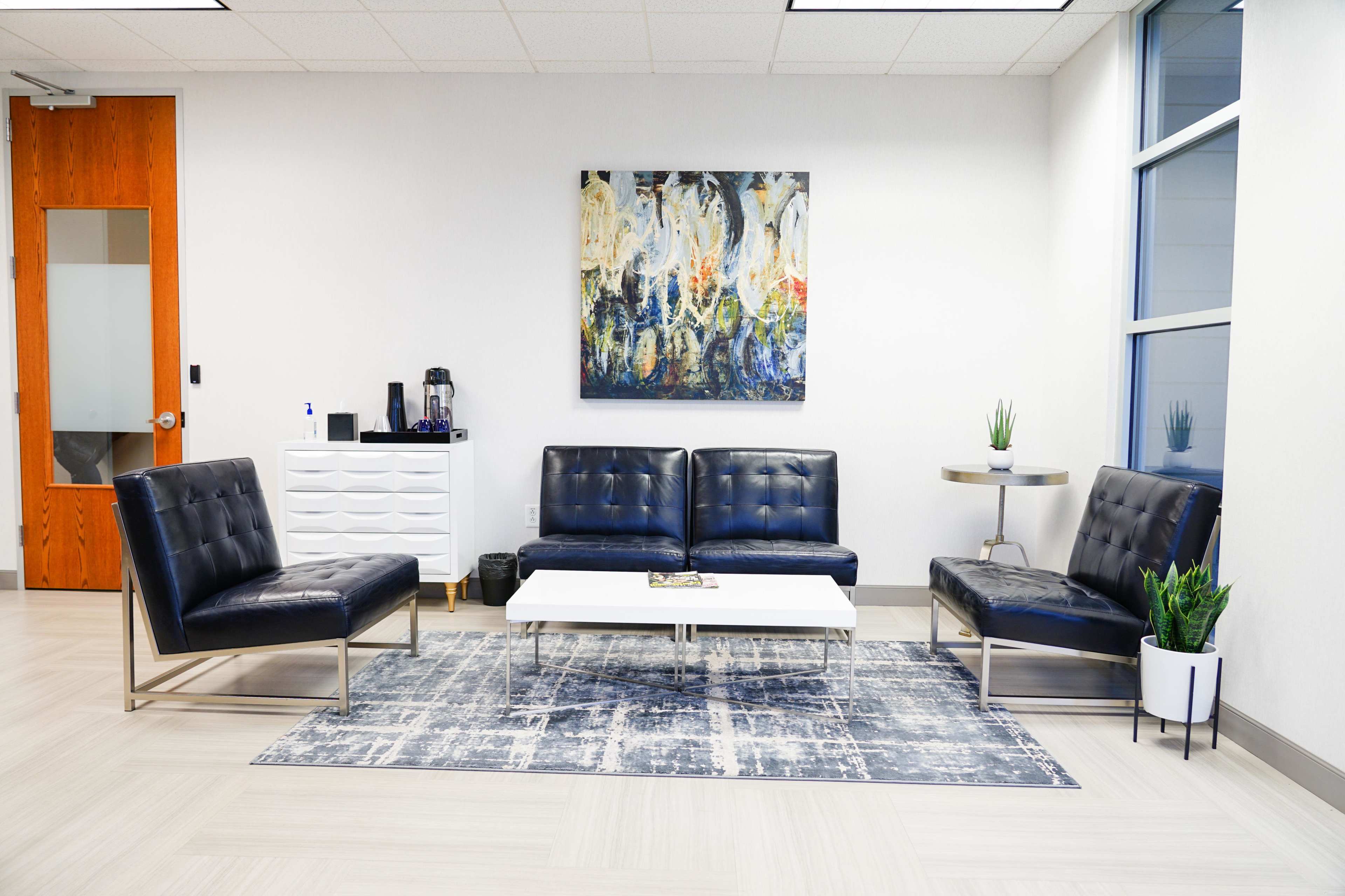 The image shows a modern waiting area featuring two black leather couches, two matching chairs, a coffee table, and a decorative wall art piece, all set on a patterned area rug.