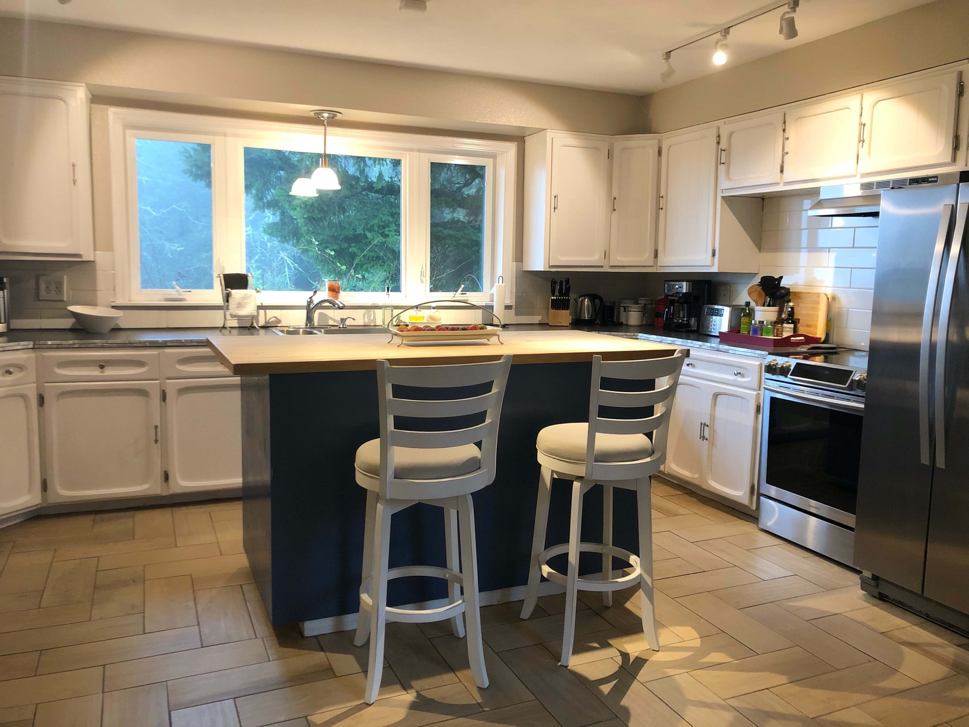The image shows a bright kitchen with white cabinetry, a central island with two tall stools, and stainless steel appliances.