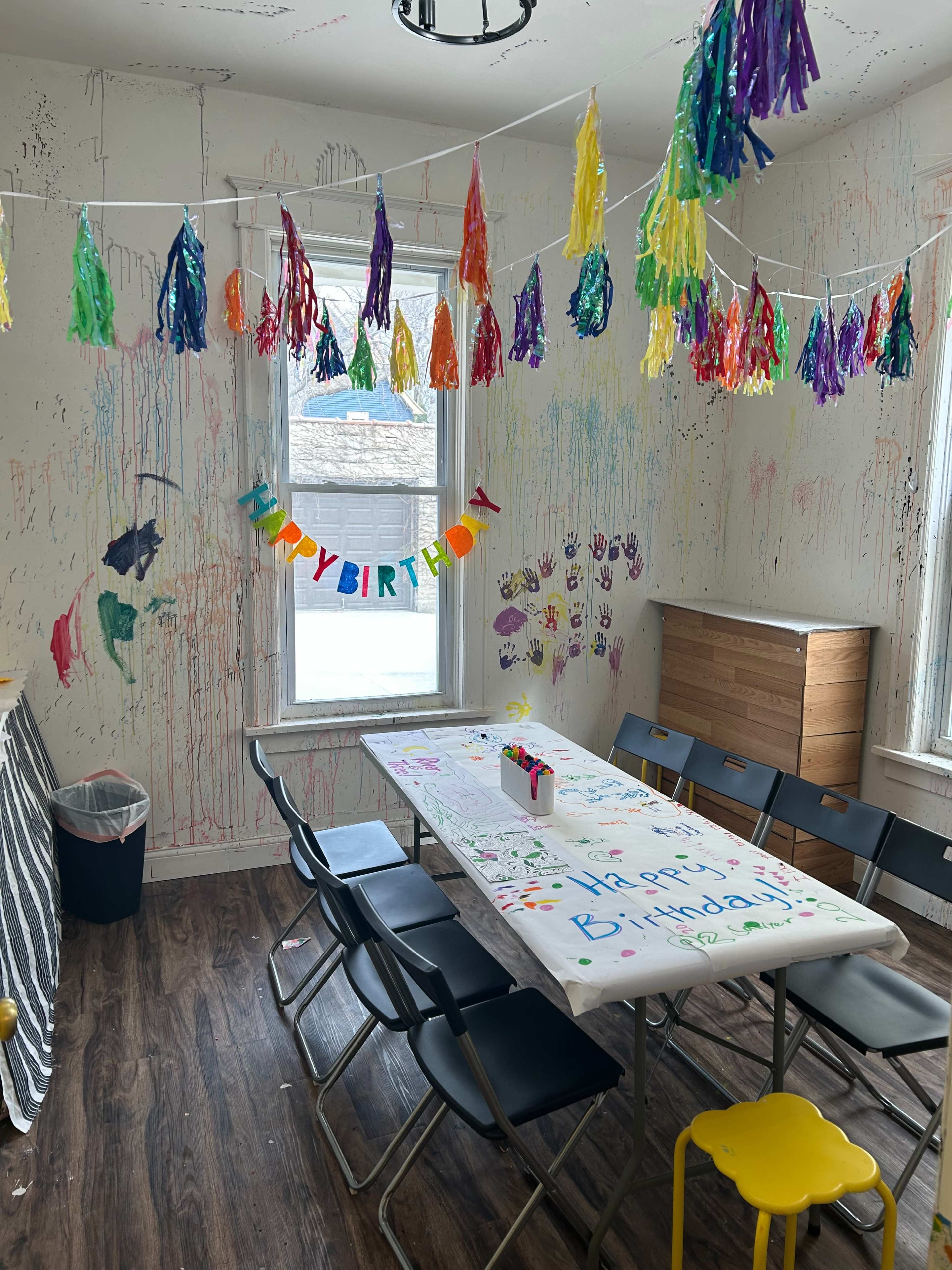 A decorated room features a table adorned with a colorful "Happy Birthday" banner and vibrant party decorations hanging from the ceiling, with walls showcasing multicolored paint streaks.