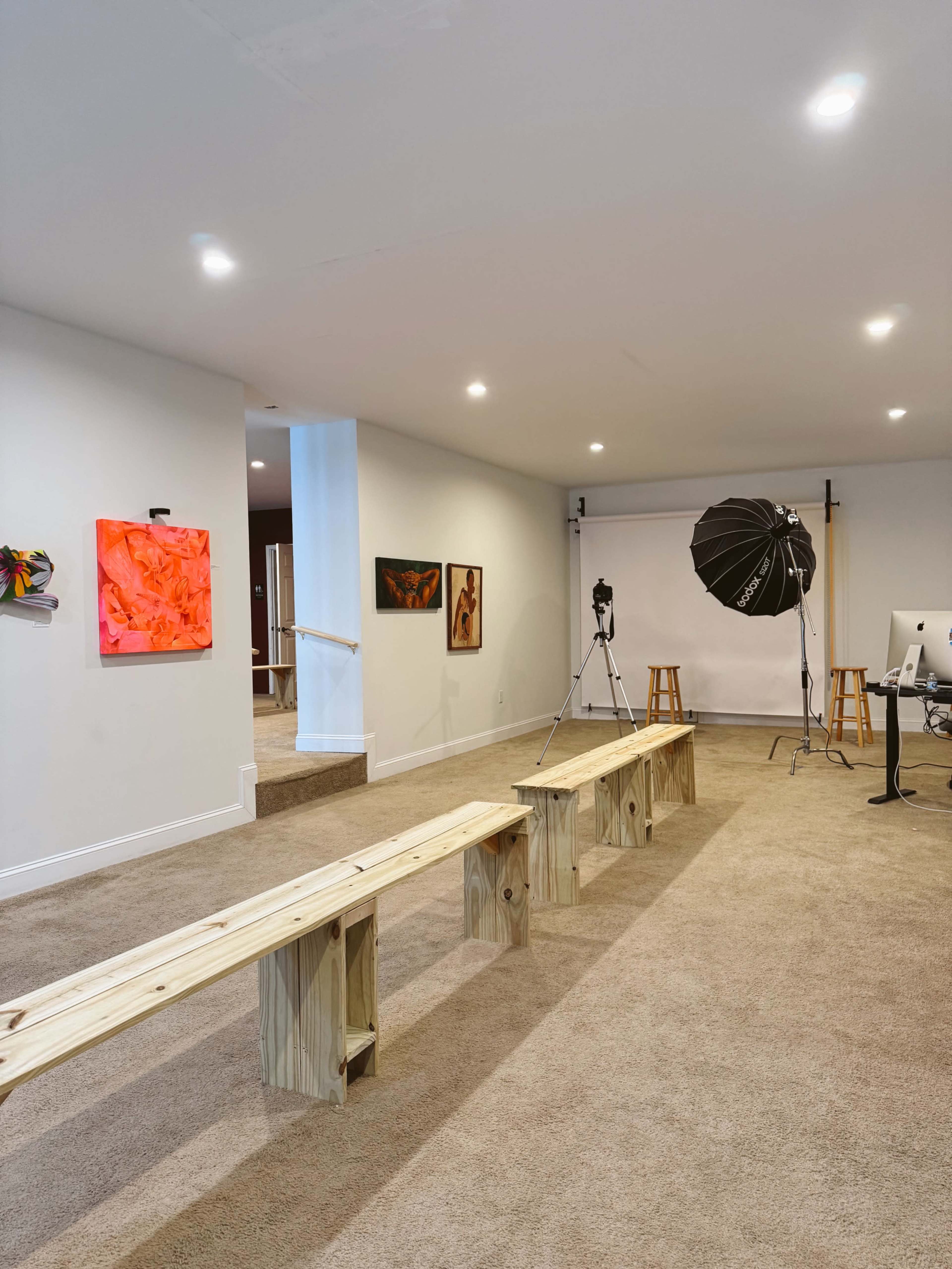 The image shows a spacious studio or gallery with wooden benches arranged in front of a photography backdrop, along with artwork on the walls and photography equipment set up in the corner.