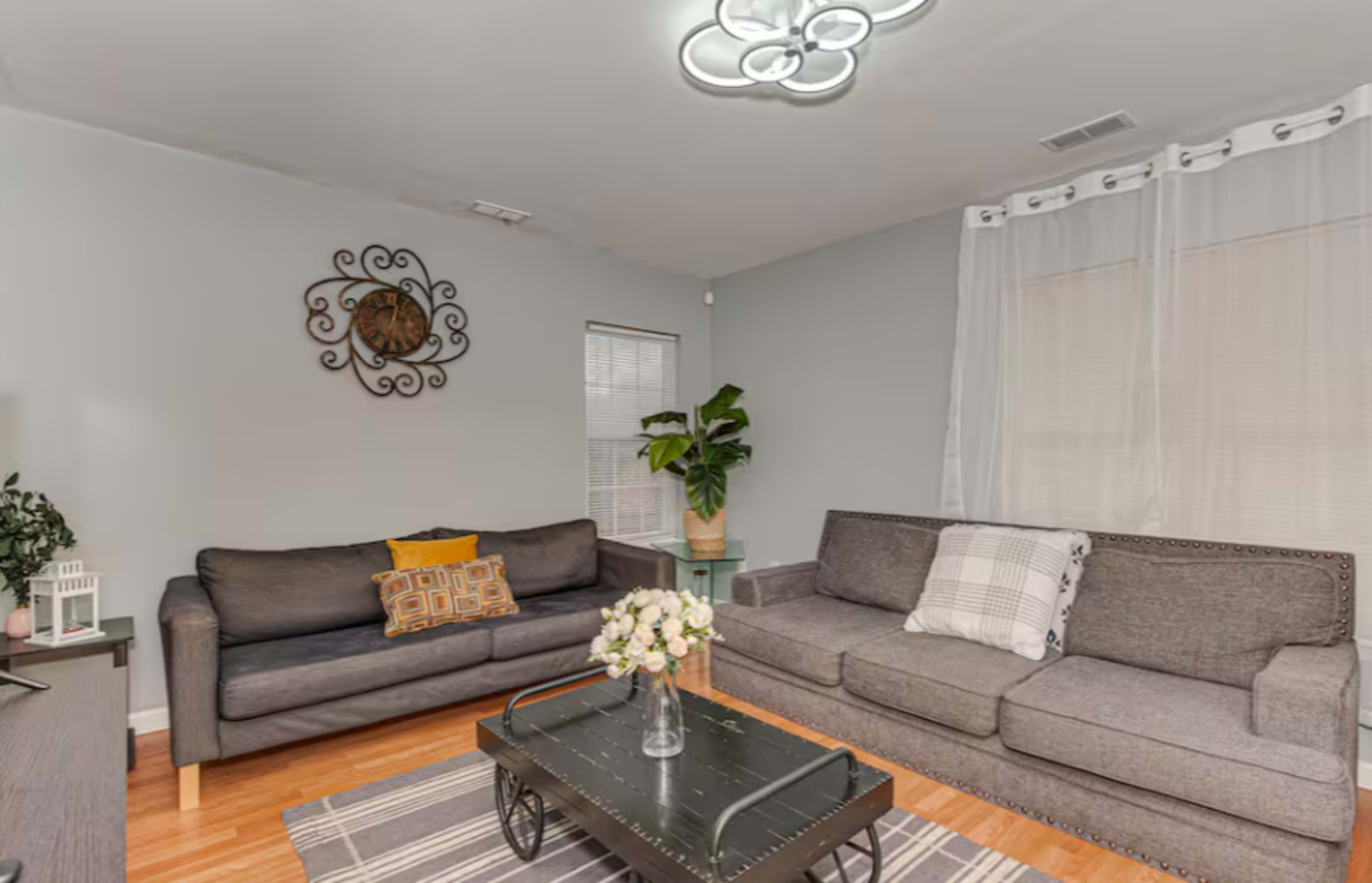 The living room features two gray sofas, a coffee table with a glass top, a small plant, and a decorative wall hanging.