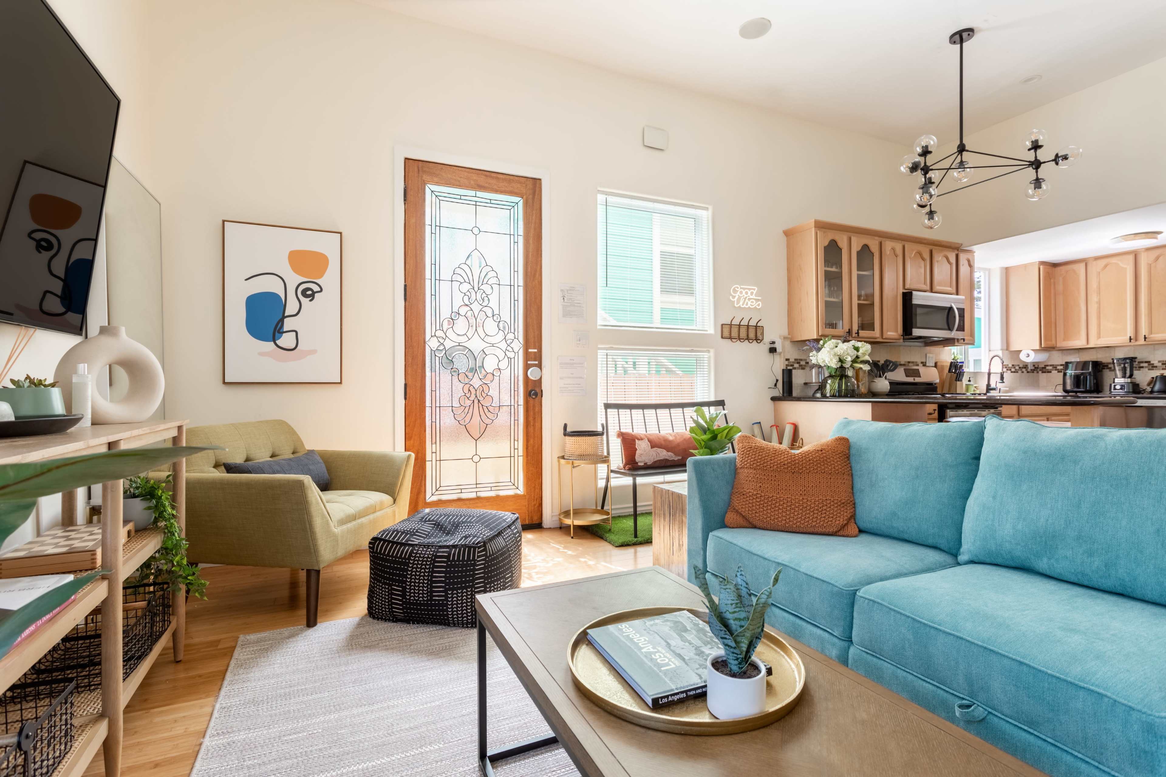 A brightly lit living room features a teal sofa, an armchair, a coffee table with a plant, and a door leading to an outdoor space.