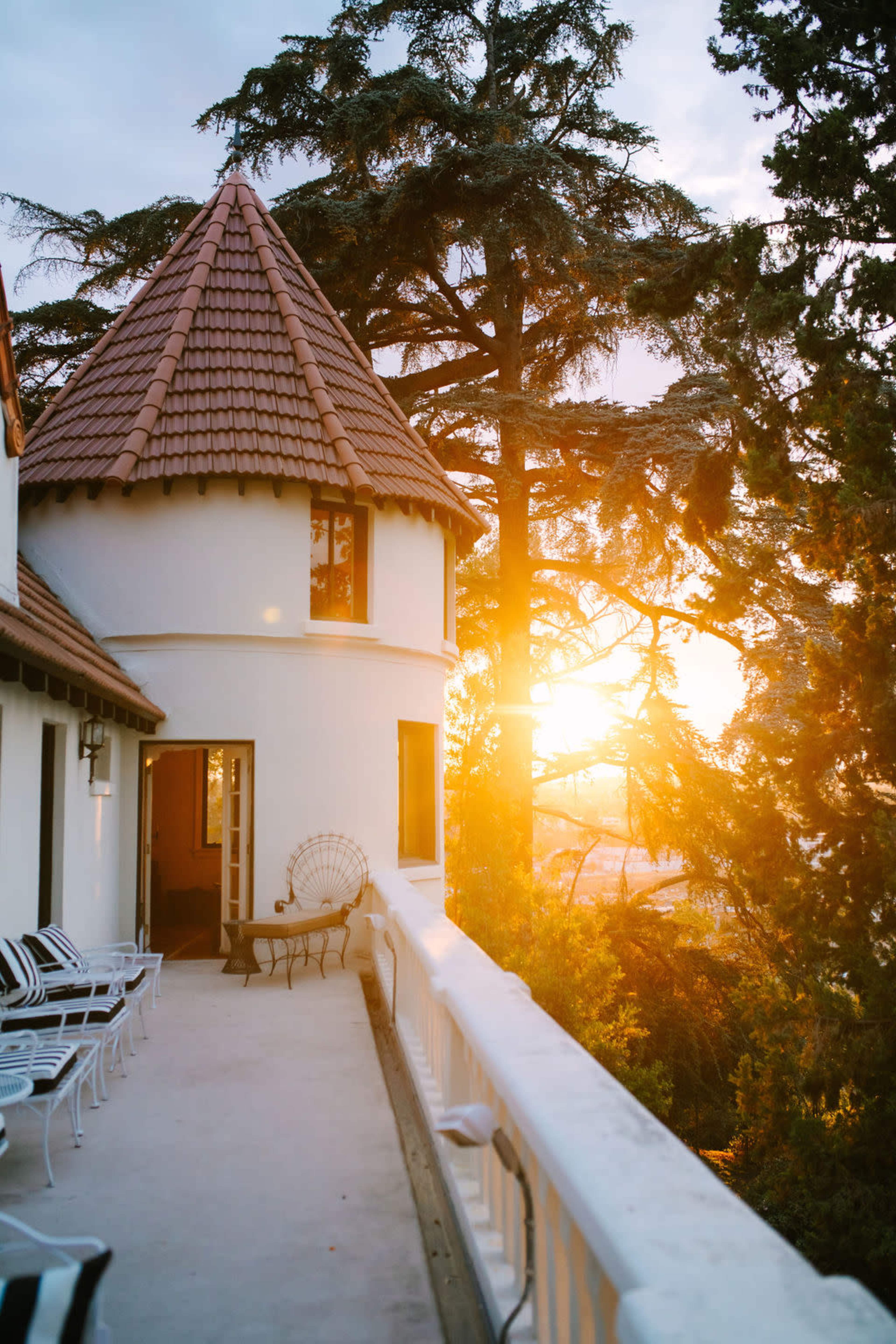The image shows a balcony of a house with a tower, surrounded by trees and illuminated by the setting sun.