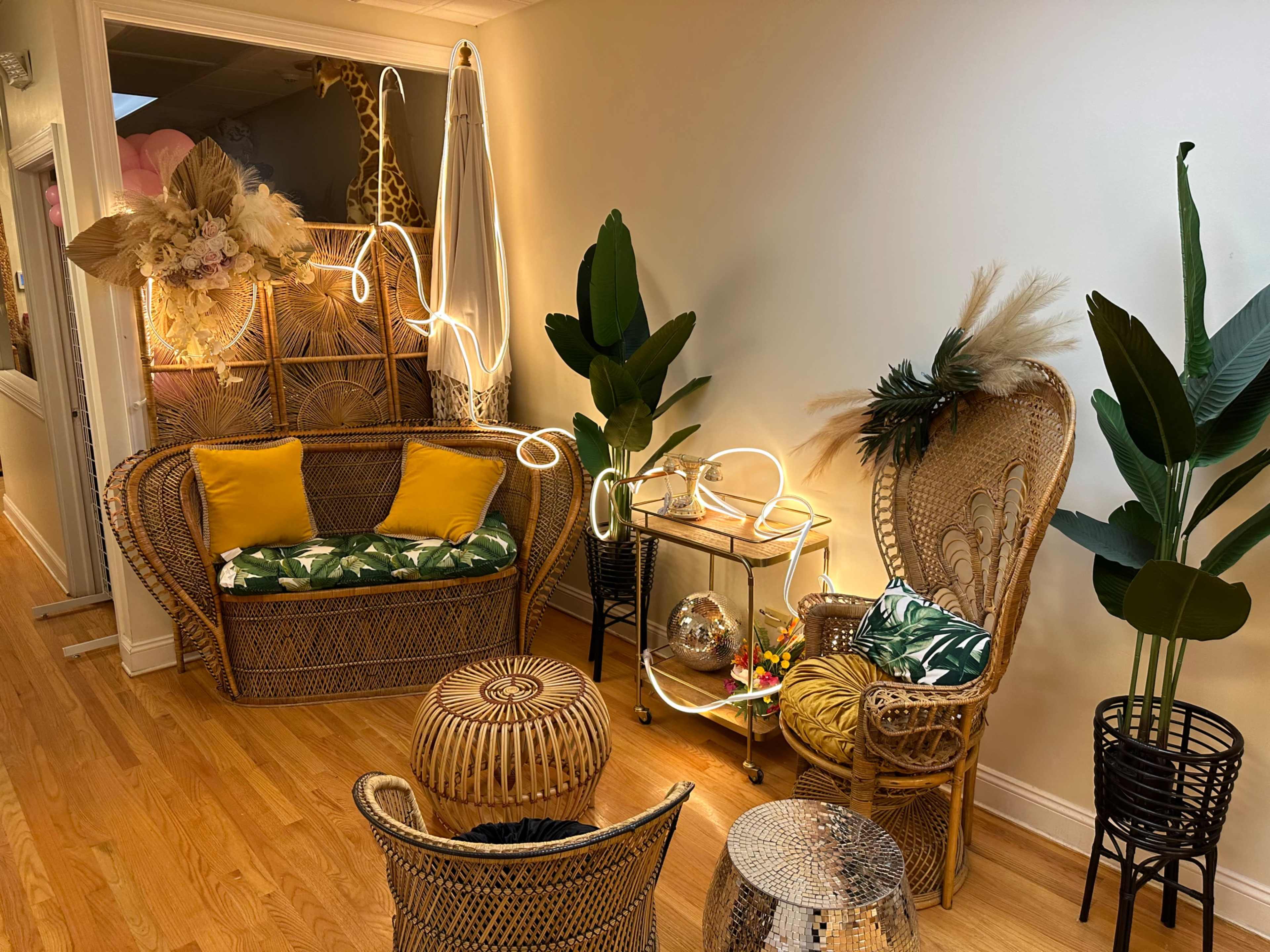 The image shows a cozy seating area with wicker furniture, colorful cushions, and decorative plants, illuminated by warm lights.