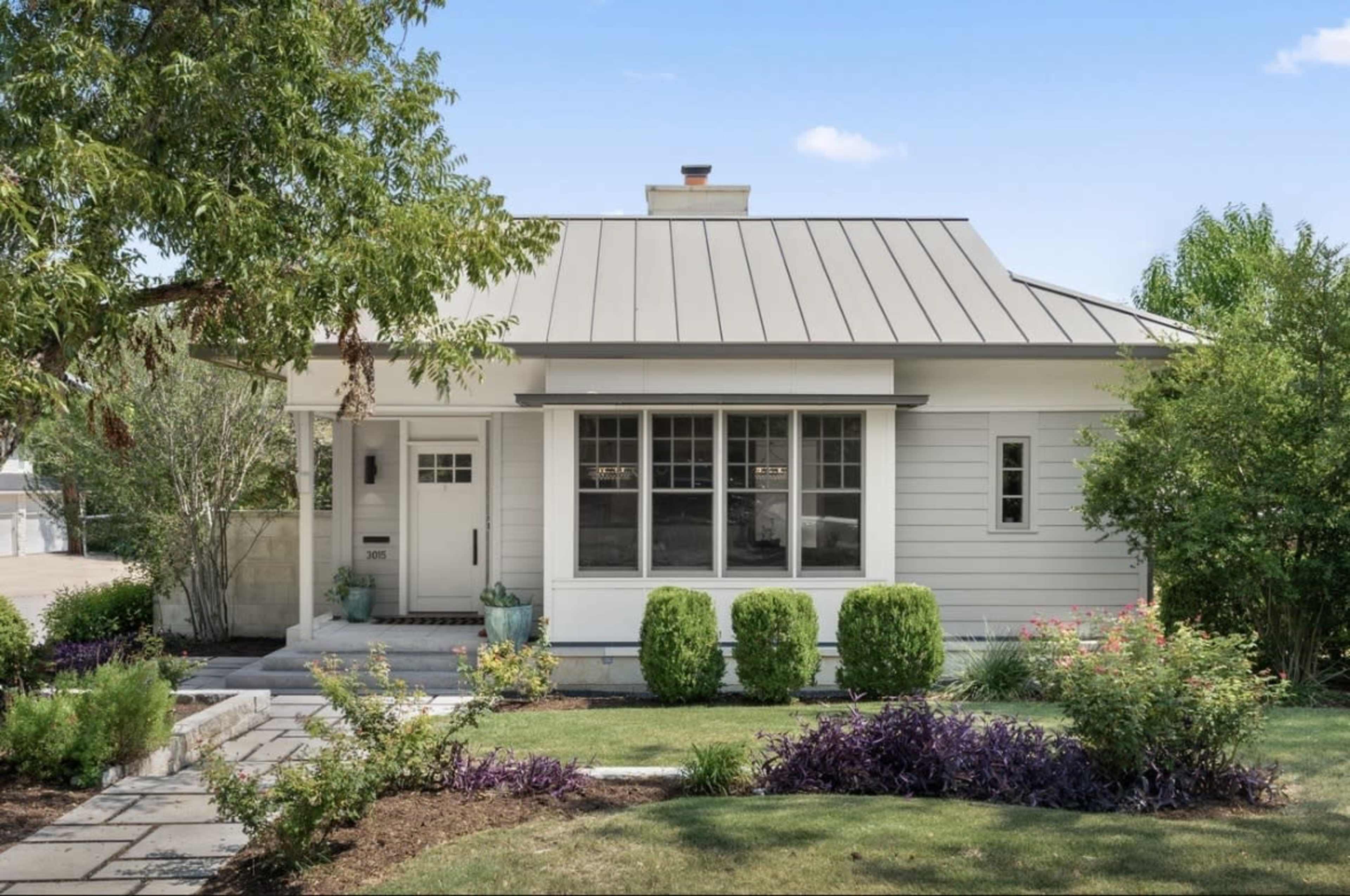 A small, modern bungalow with a metal roof features a front porch, large windows, and landscaped gardens.