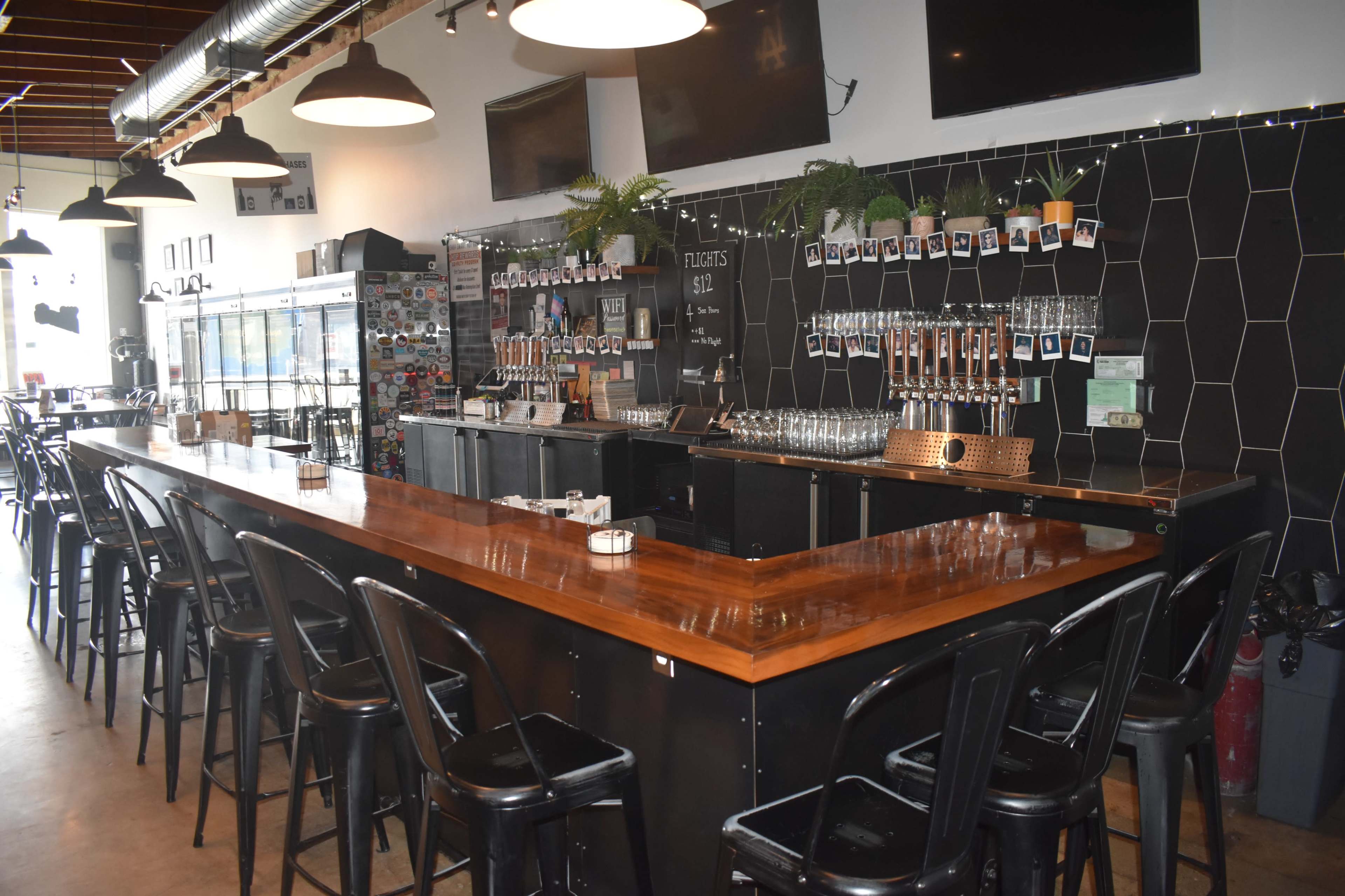 The image shows a modern bar with a wooden countertop, metal stools, and a row of beer taps against a black hexagonal-patterned wall.