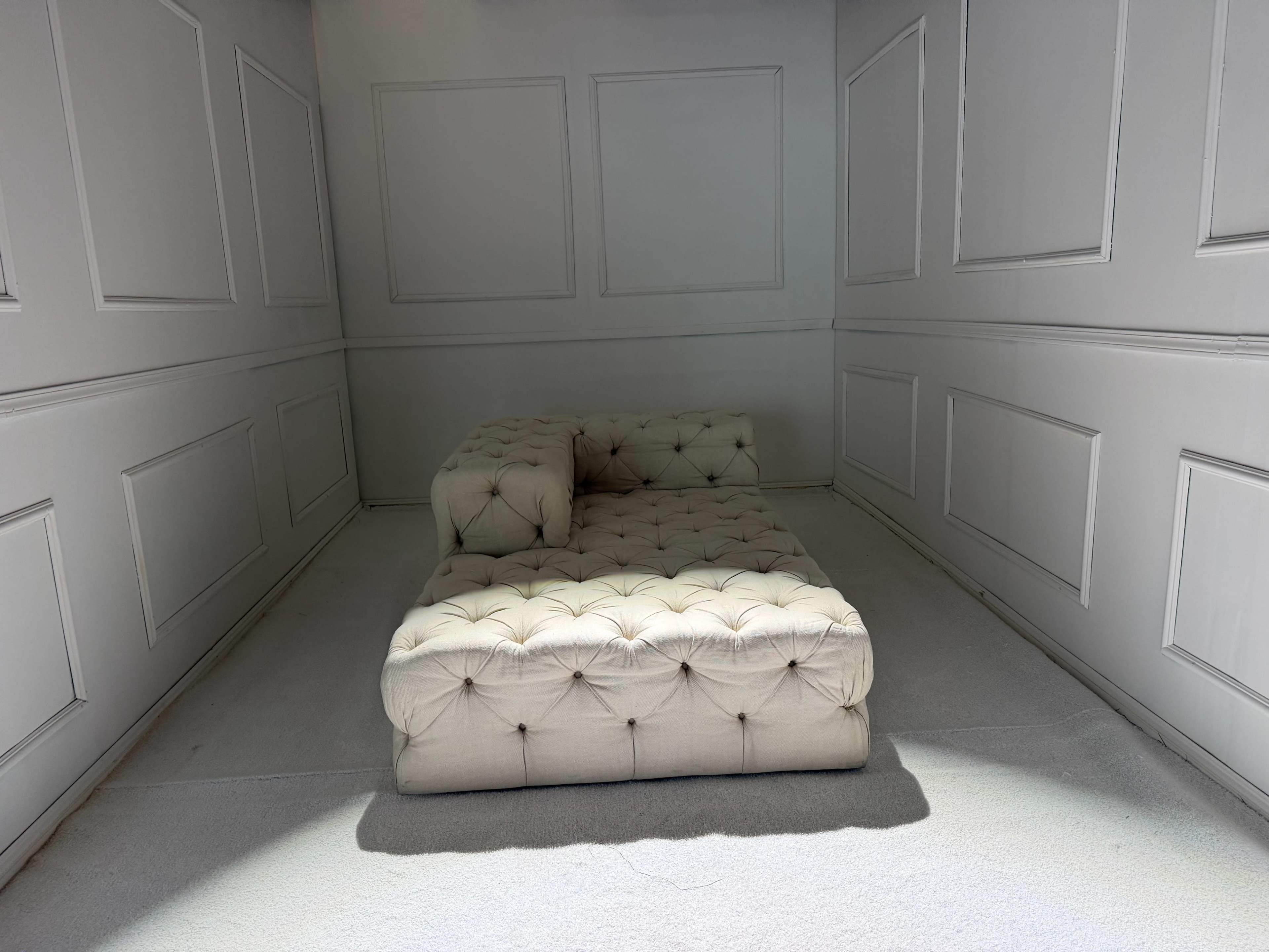 The image shows a tufted, light-colored sofa positioned in an empty, minimally decorated room with paneled walls.